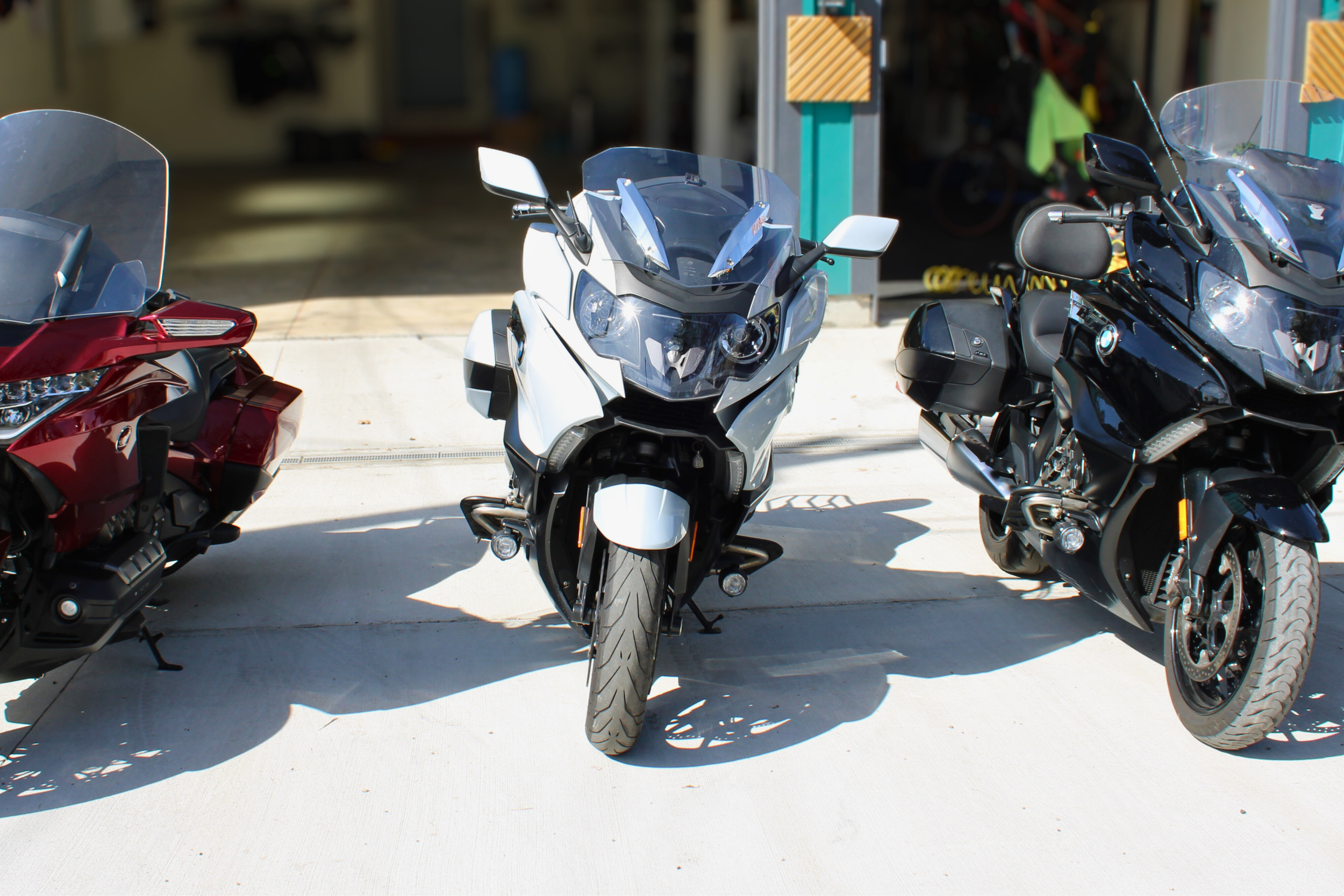 Three motorcycles parked side by side in front of a garage, with the middle one being a white BMW motorcycle and the other two being black and red BMW motorcycles.