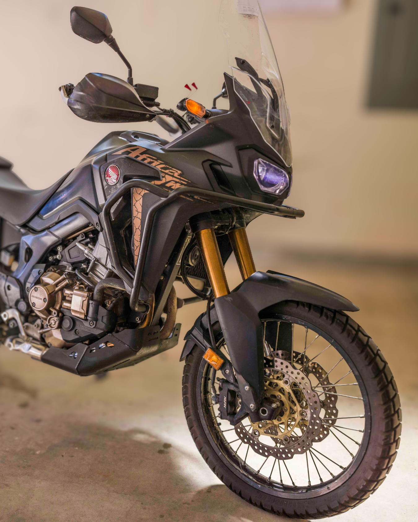 Black Honda Africa Twin adventure motorcycle parked on a sidewalk with other motorcycles in the background, trees with autumn leaves, and sunlight creating a lens flare.