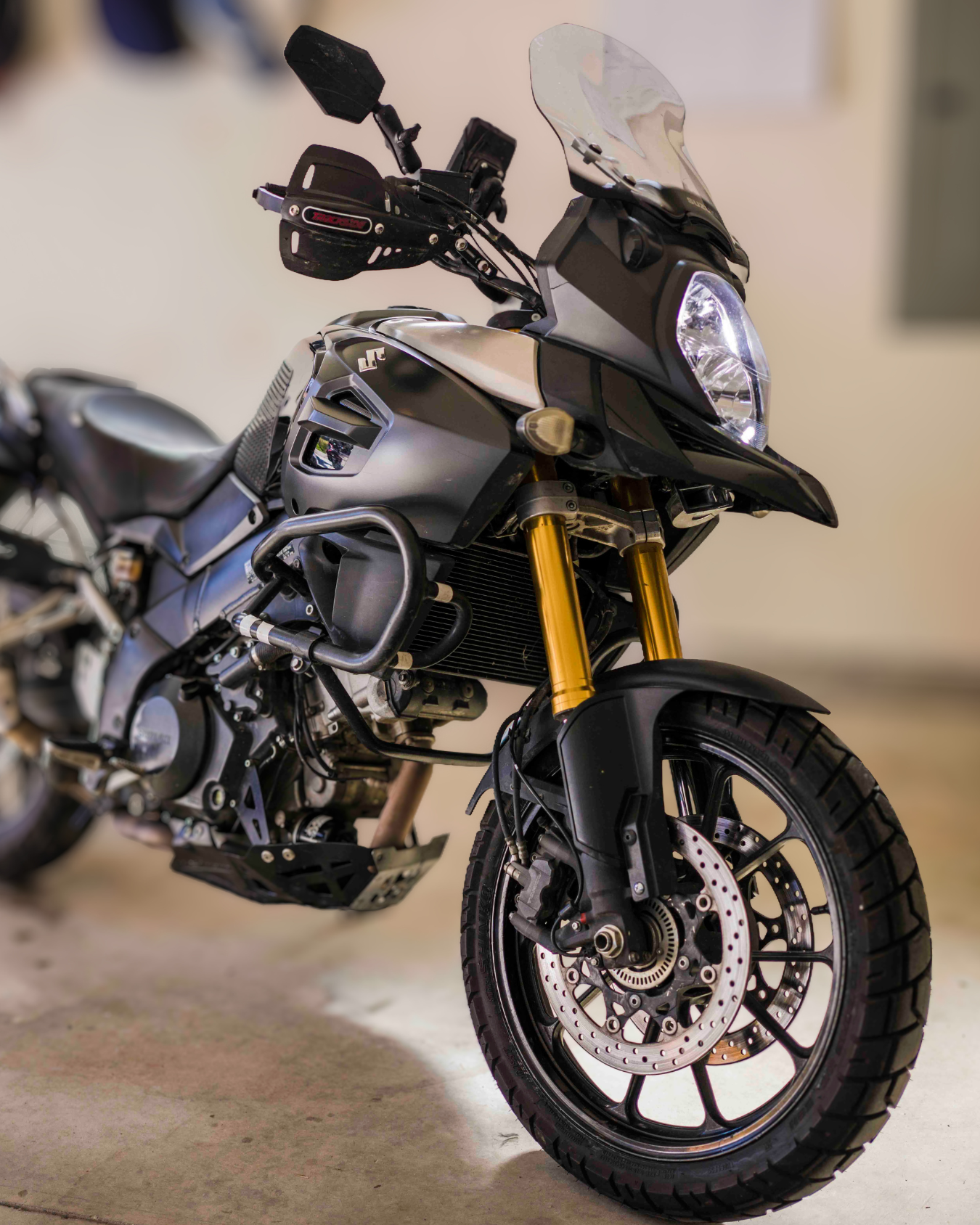 A black and silver Suzuki adventure motorcycle with a front disc brake, gold front forks, and a clear windshield, parked indoors.