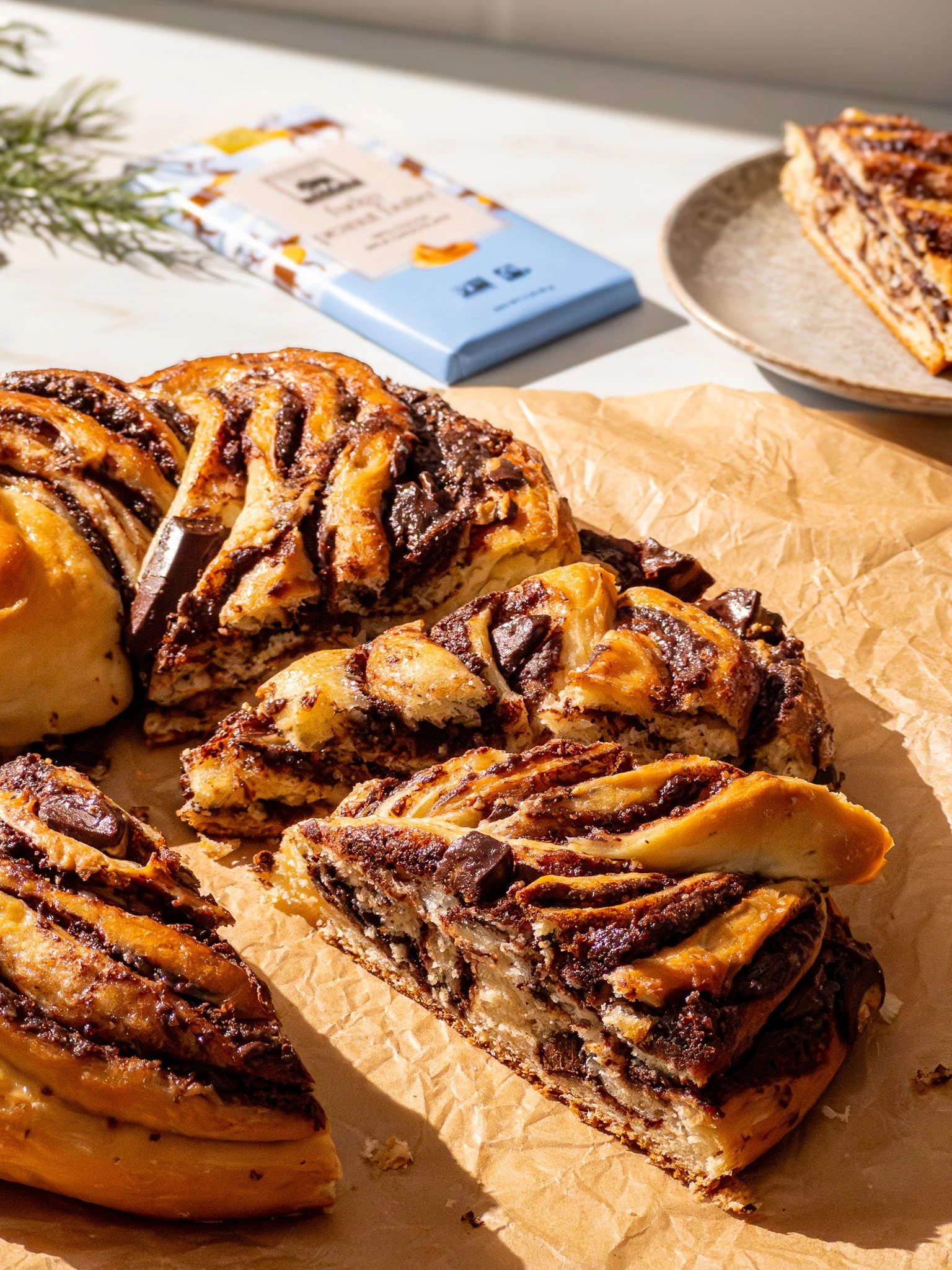 Sliced chocolate chip and cookie dough swirl cake on parchment paper, with chocolate chunks visible inside, and a chocolate bar and plate with a slice of the cake in the background.