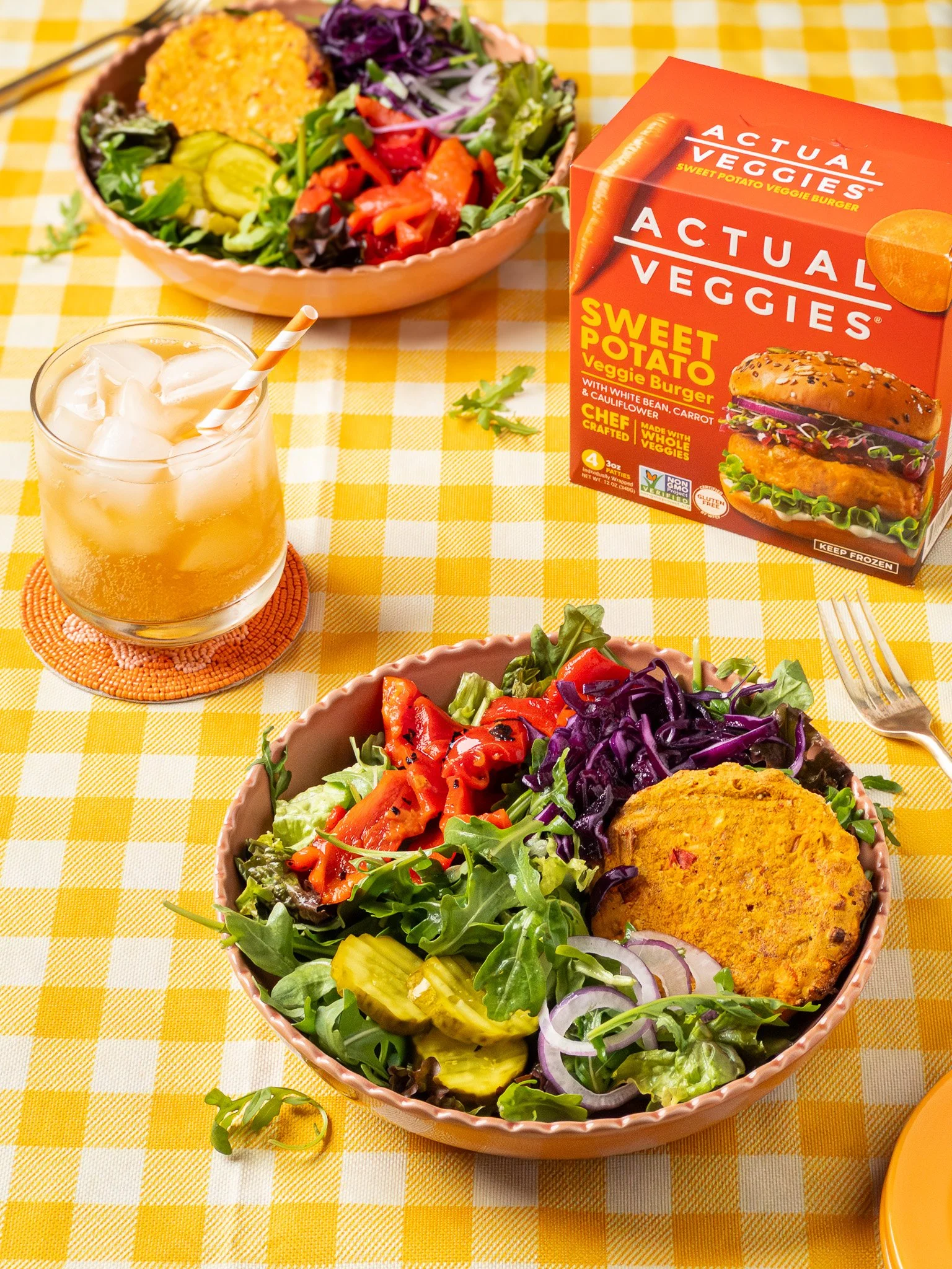 A green salad with shredded red cabbage, sliced red tomatoes, sliced onions, cucumber slices, and a veggie burger patty in a pink bowl, with a glass of iced beverage and a box of vegan veggie burger on a yellow checkered tablecloth.