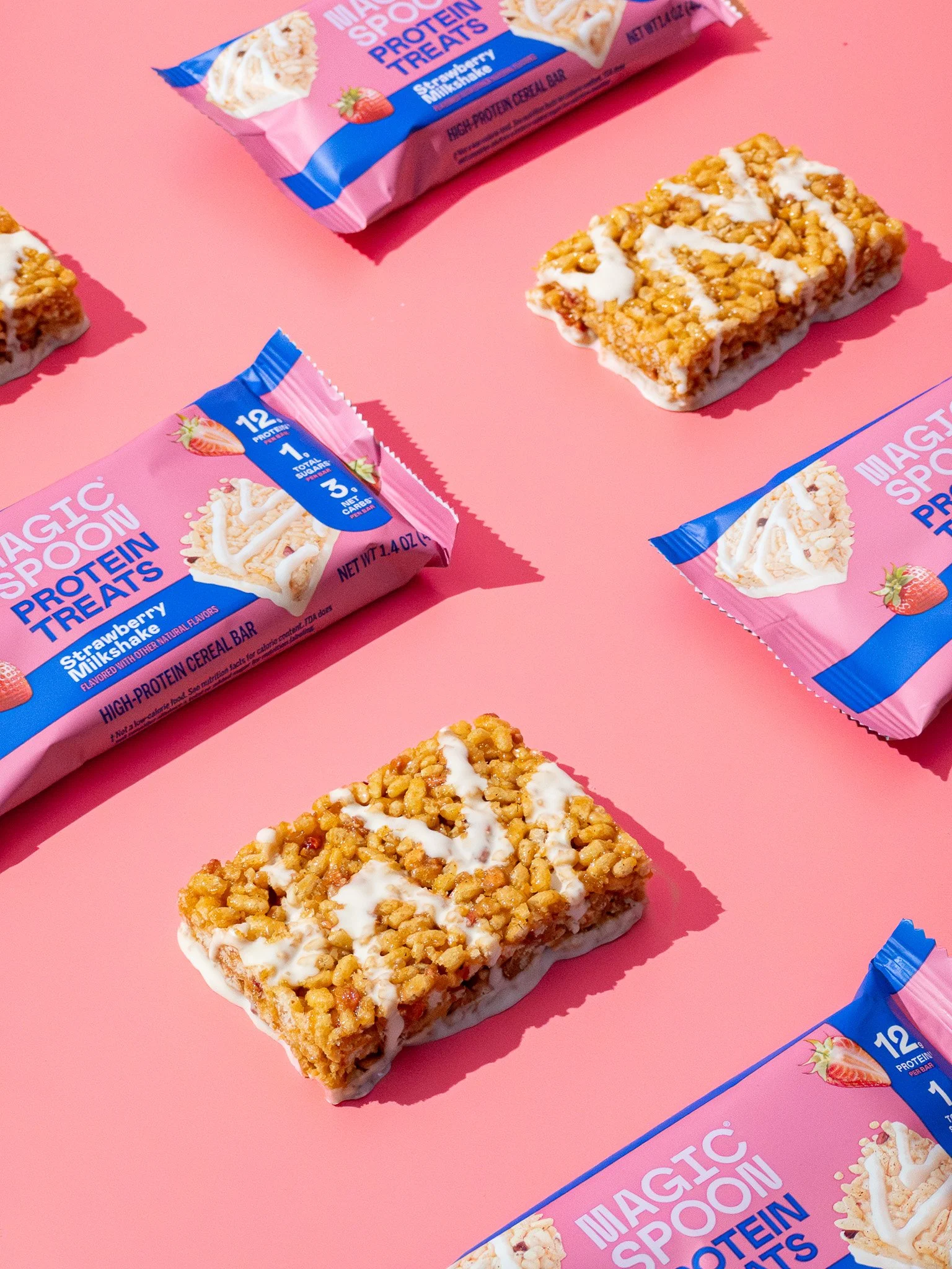 Multiple packages of Magic Spoon protein treats in strawberry milkshake flavor, placed on a pink background alongside rice crispy treats with white icing.