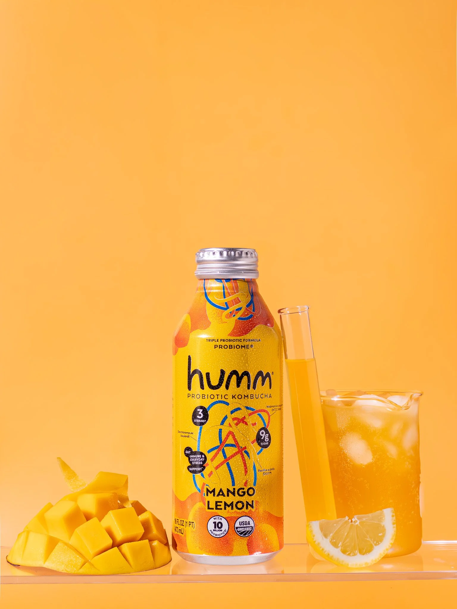 A bottle of Humm Mango Lemon probiotic kombucha, a glass of cold kombucha with ice and lemon slices, a test tube with kombucha, and a sliced mango, all set against a yellow background.