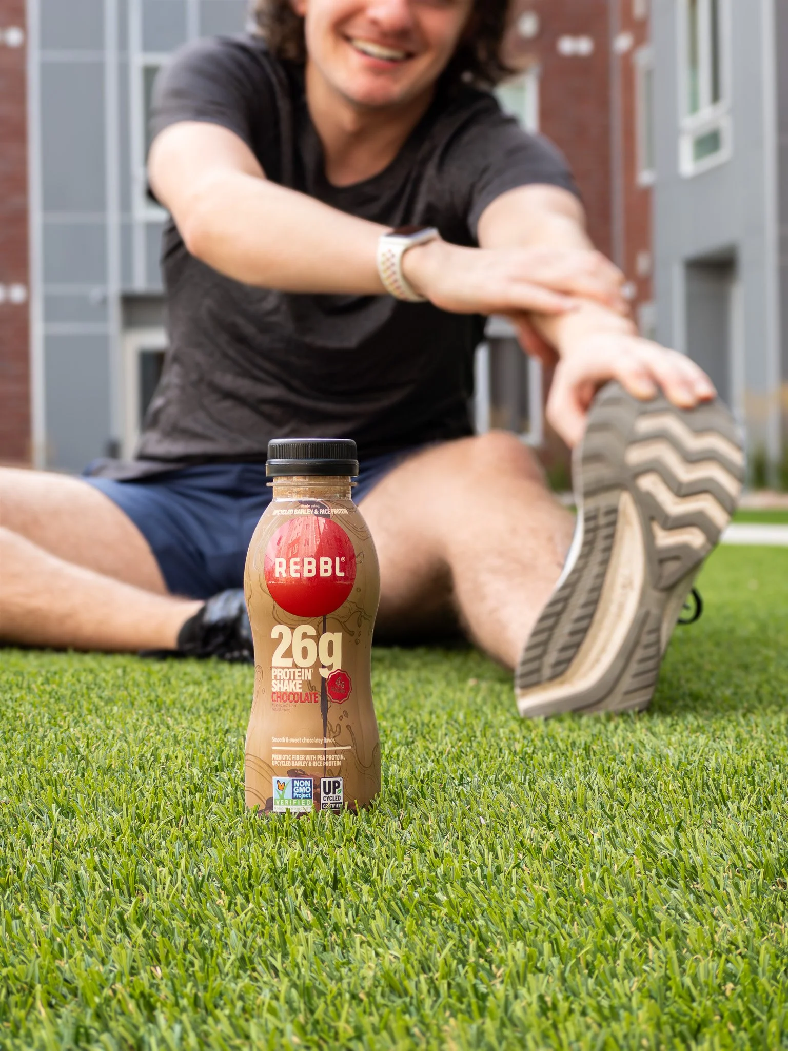 A man stretching outdoors on grassy ground with a chocolate-flavored protein shake bottle in the foreground.