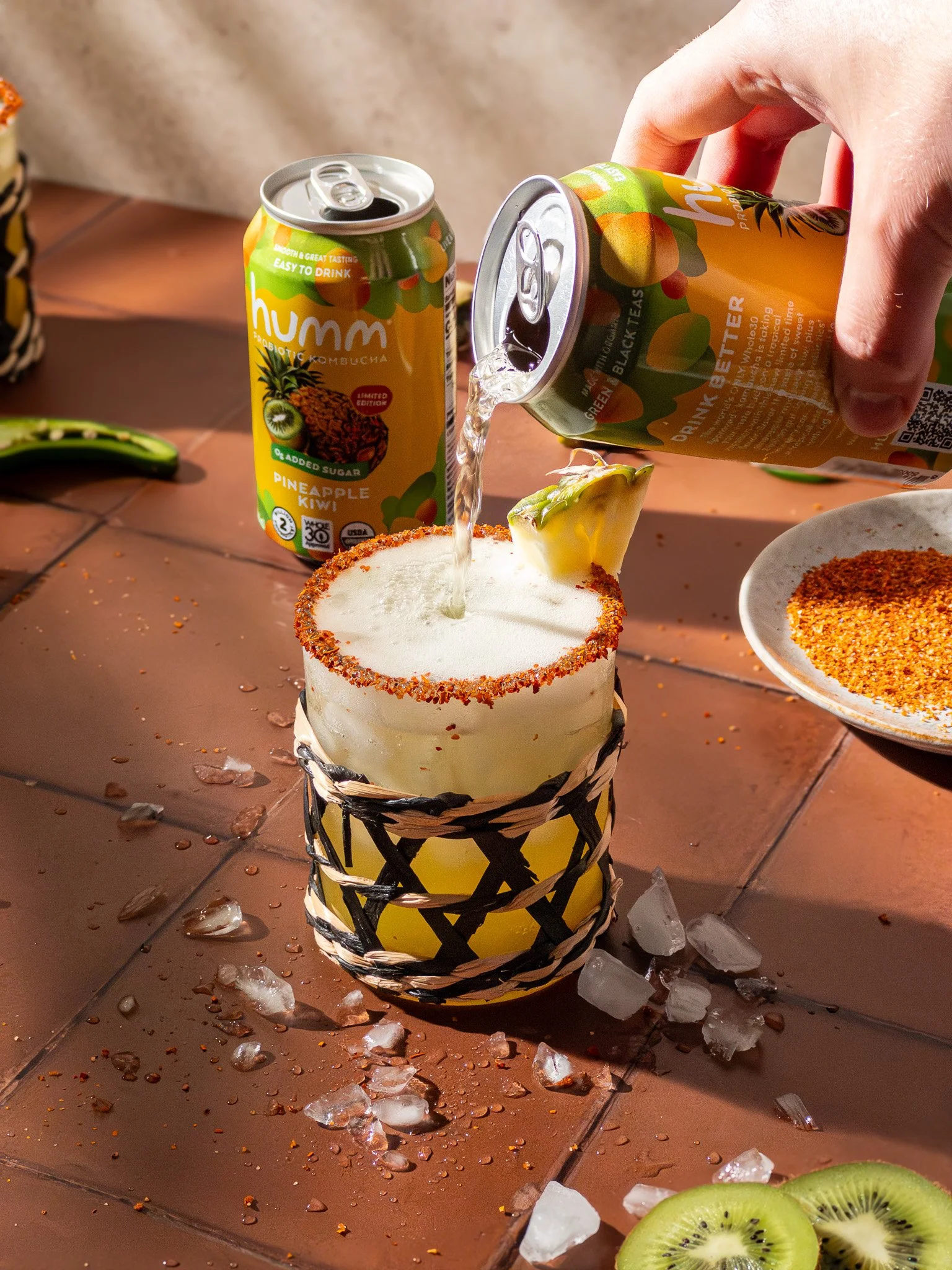 A hand pouring sparkling water into a layered cocktail glass with a yellow and black woven design, garnished with pineapple. There are cans of kombucha and a plate of red seasoning on a brown tiled table, with scattered ice and kiwi slices nearby.