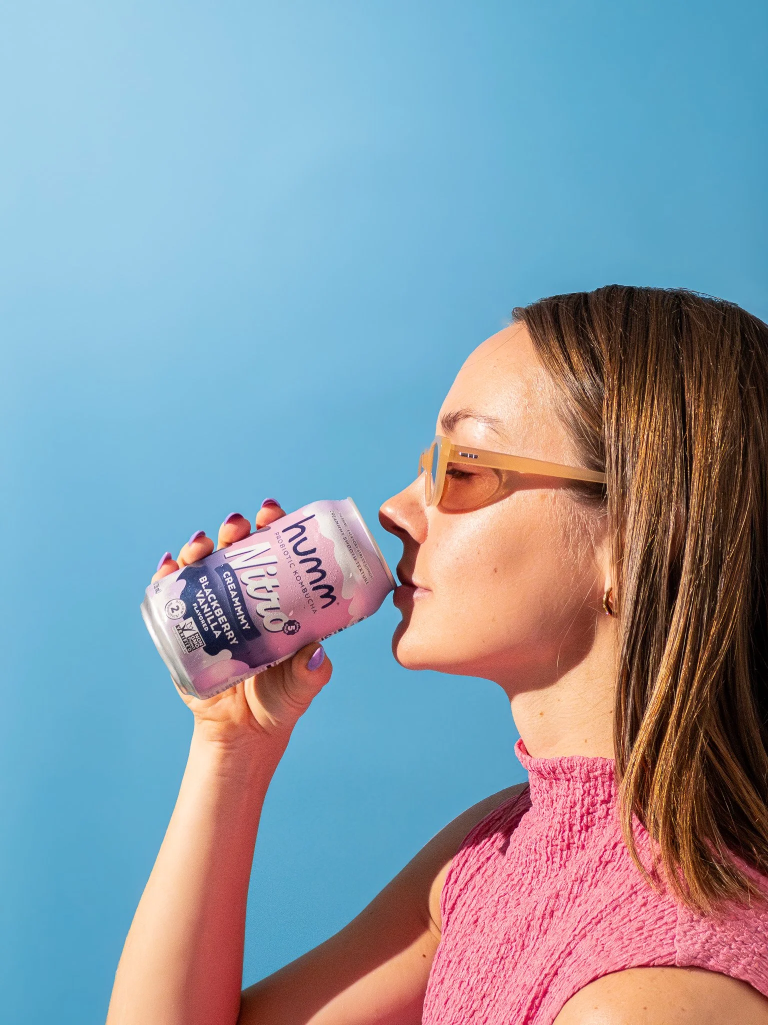 A woman wearing sunglasses and a pink sleeveless top is holding a can of Humm Vita probiotic beverage close to her mouth against a bright blue sky background.