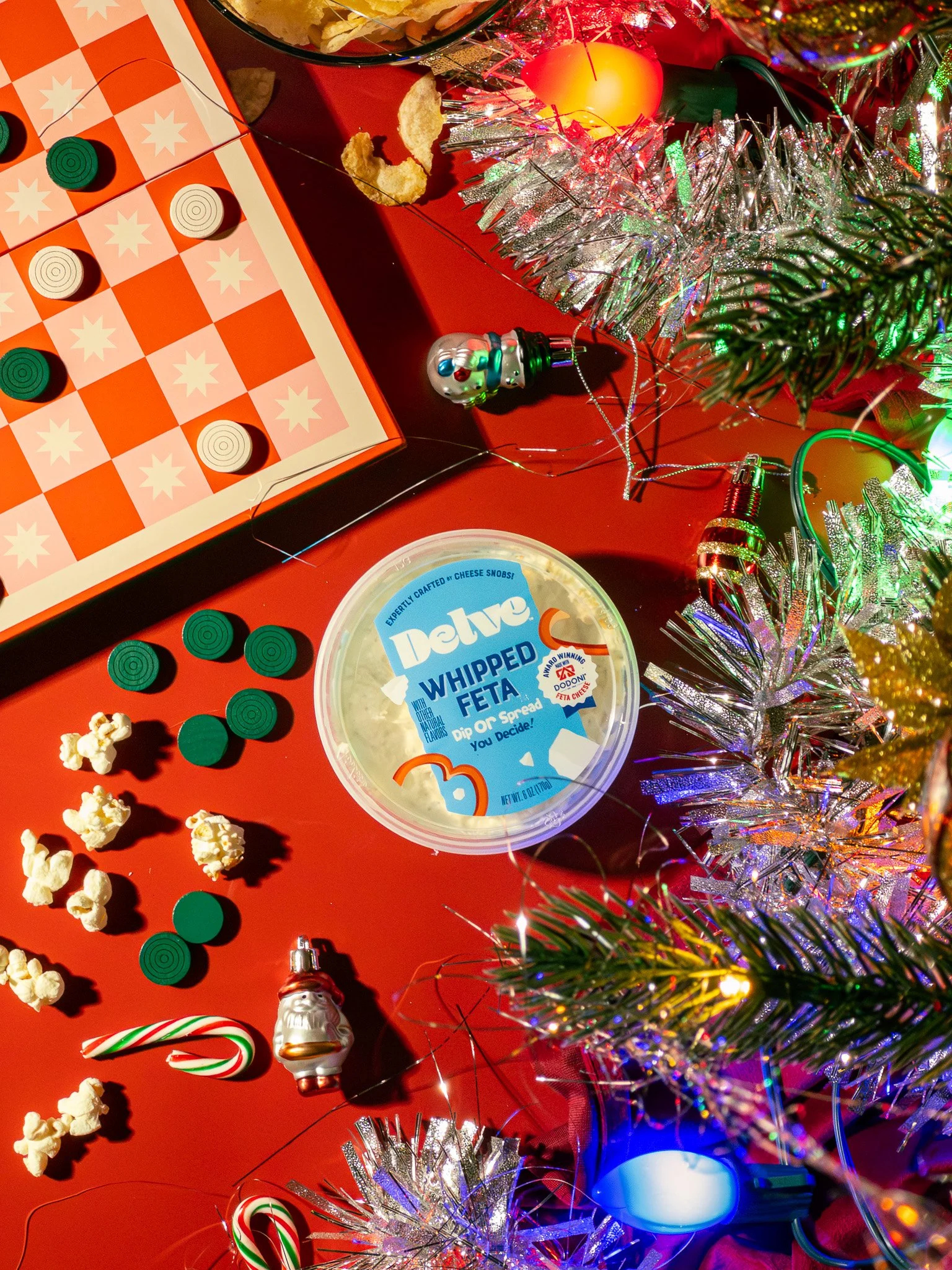 A festive holiday scene featuring a red table with Christmas decorations, a tub of Delve whipped feta cheese, popcorn, candy canes, a snowman ornament, and a colorful checkers game. There are tinsel, string lights, and a flickering candle adding to t