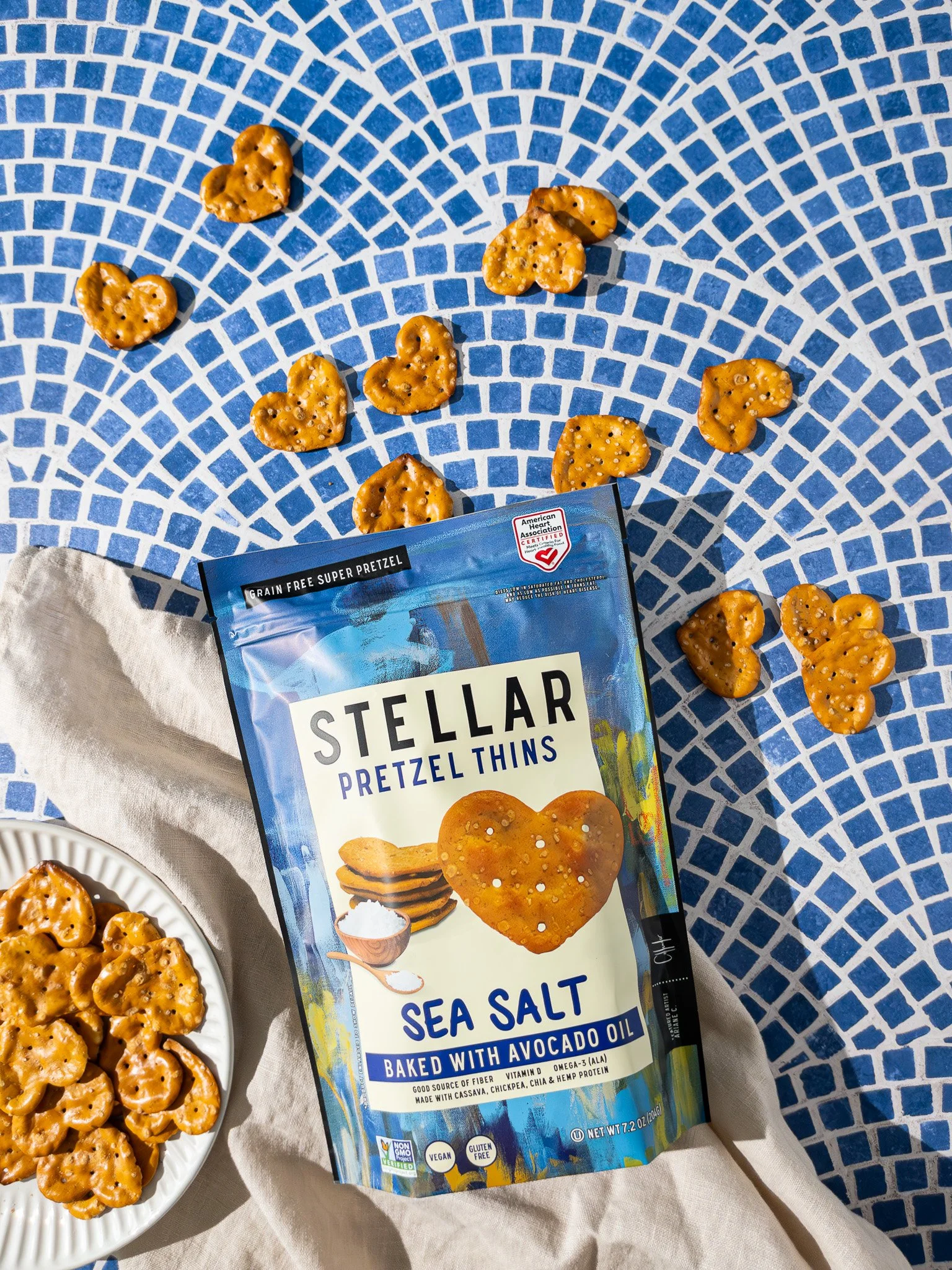 A bag of Stellar Pretzel Thins Sea Salt flavor with a plate of pretzel thins beside it, on a white and blue mosaic table with scattered pretzel thins around.