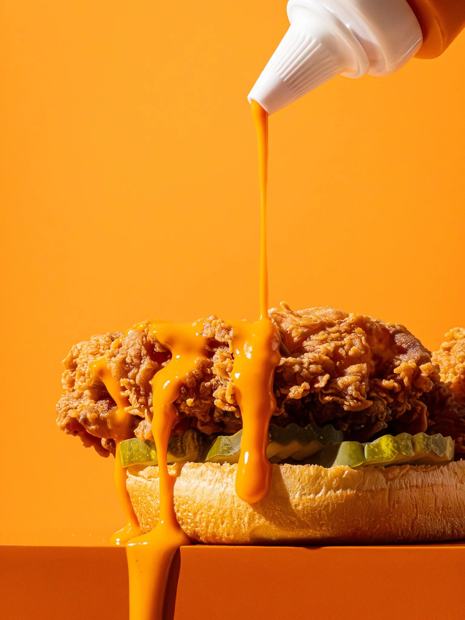 Fried chicken sandwich with pickles on a bun, drizzled with bright orange sauce, with a squeeze bottle dispensing more sauce over it, against an orange background.