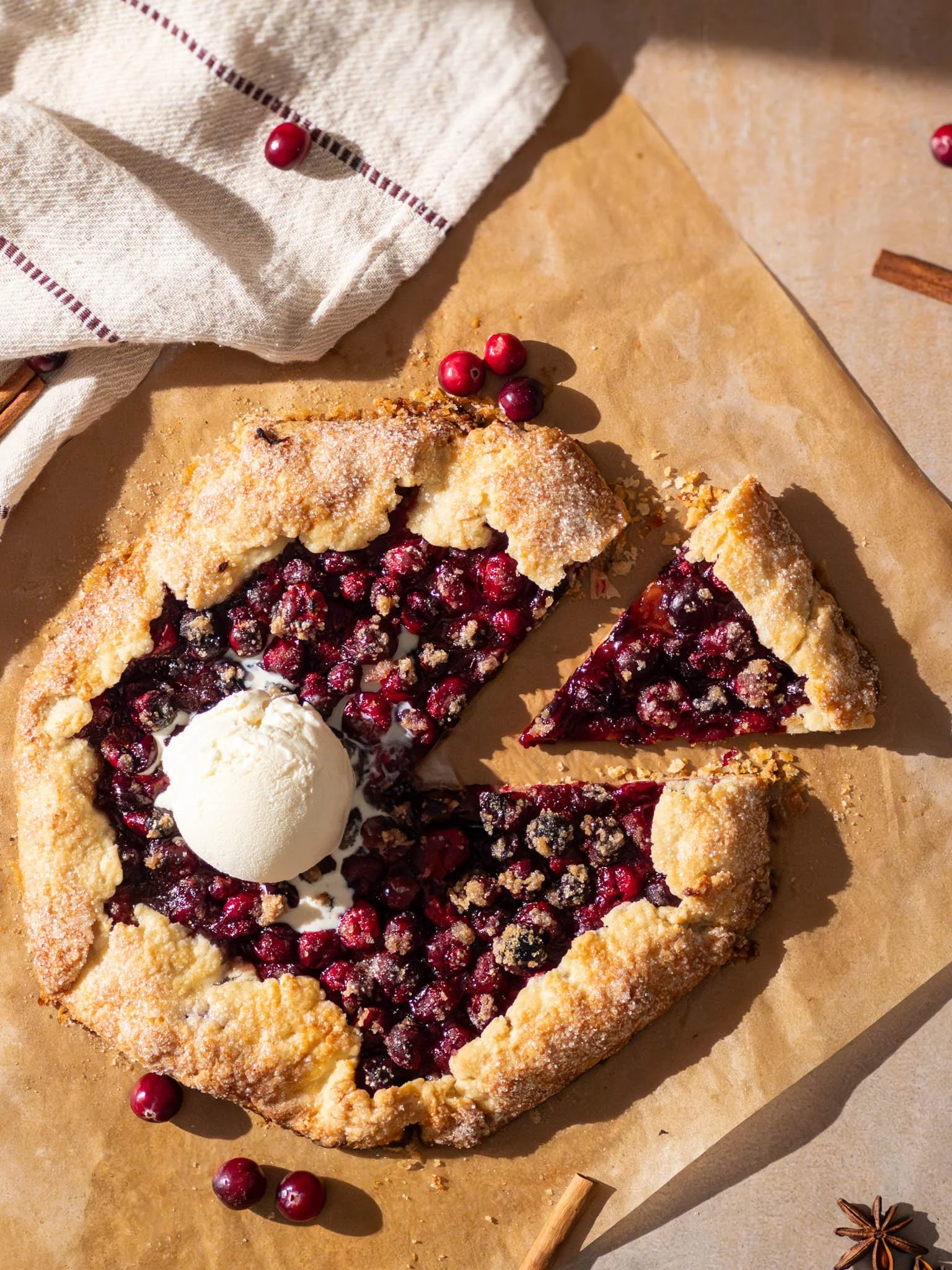 A slice of cherry galette with a scoop of vanilla ice cream on top, on parchment paper with scattered cranberries and spices.