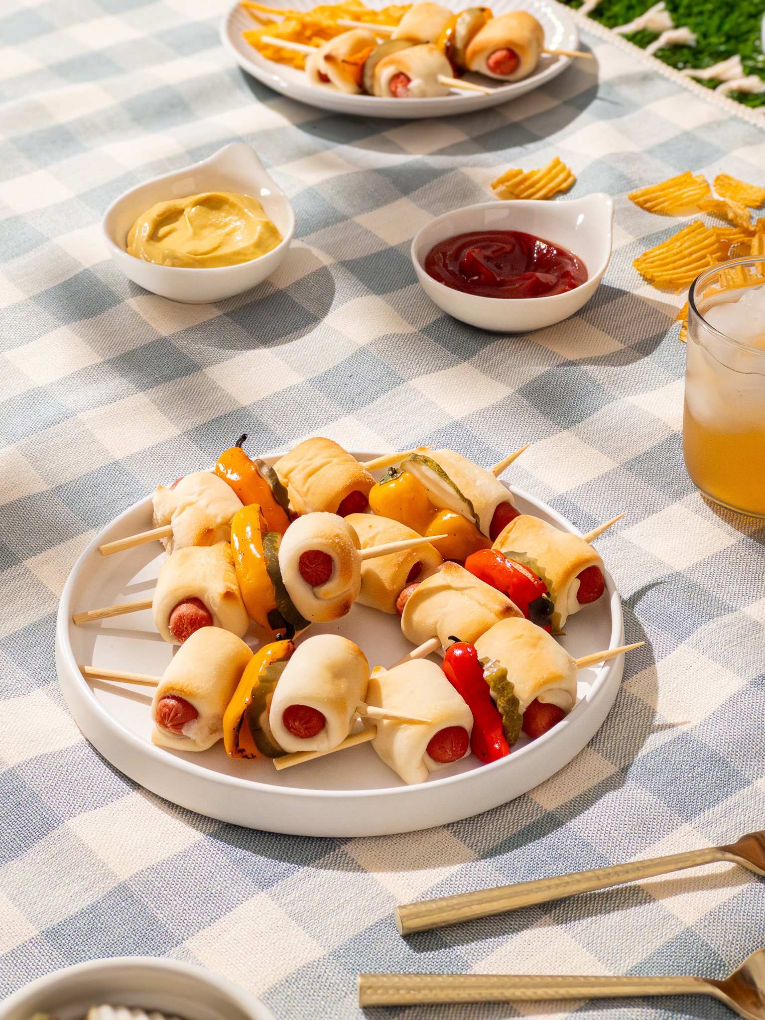 Plate of hot dogs wrapped in dough with peppers, served on skewers, on a checkered tablecloth with condiments and side dishes.