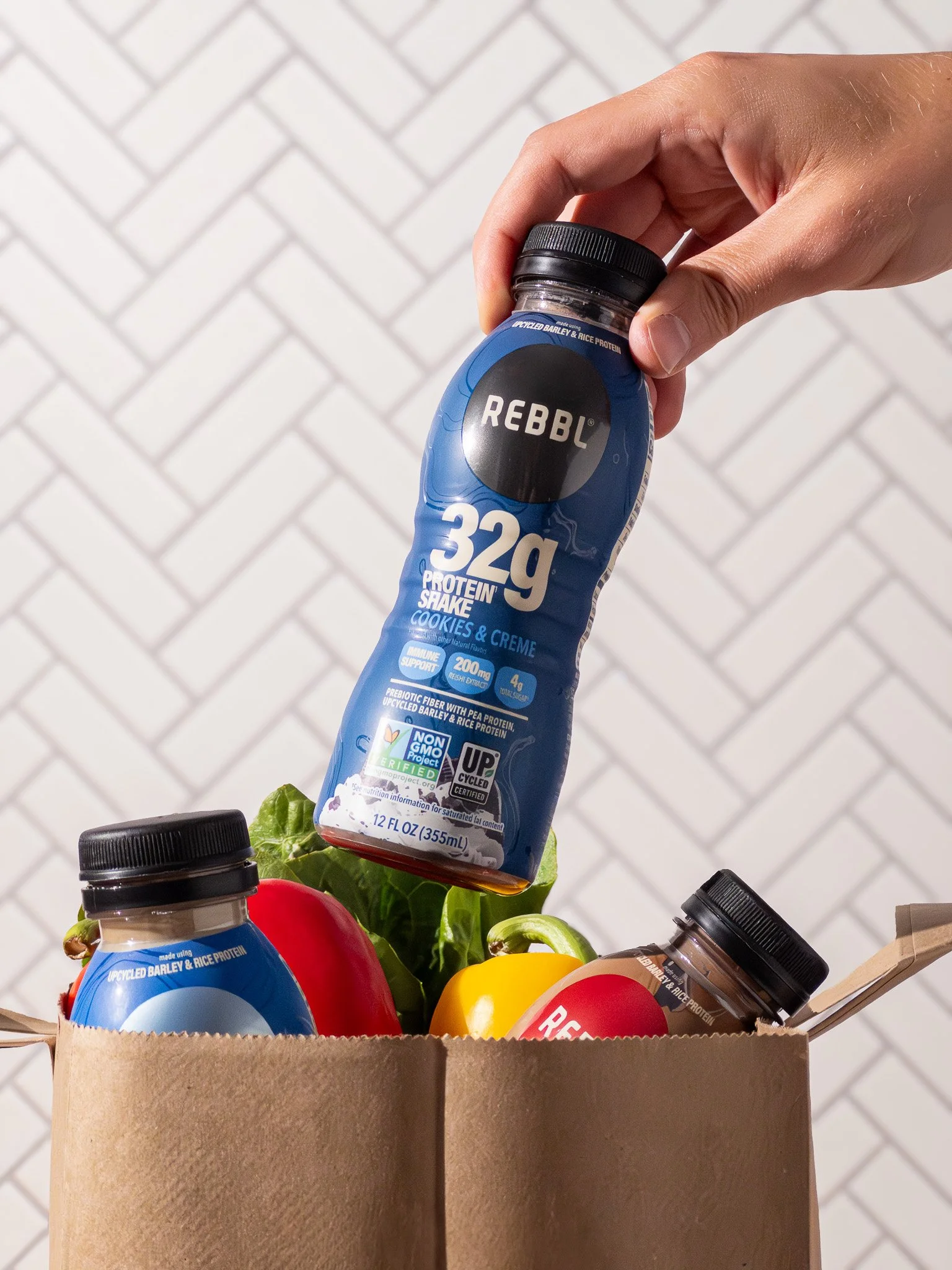 Hand holding a blue bottle of REBBL 32g protein shake with cookies and creme flavor, above a grocery bag filled with bell peppers and other bottles.