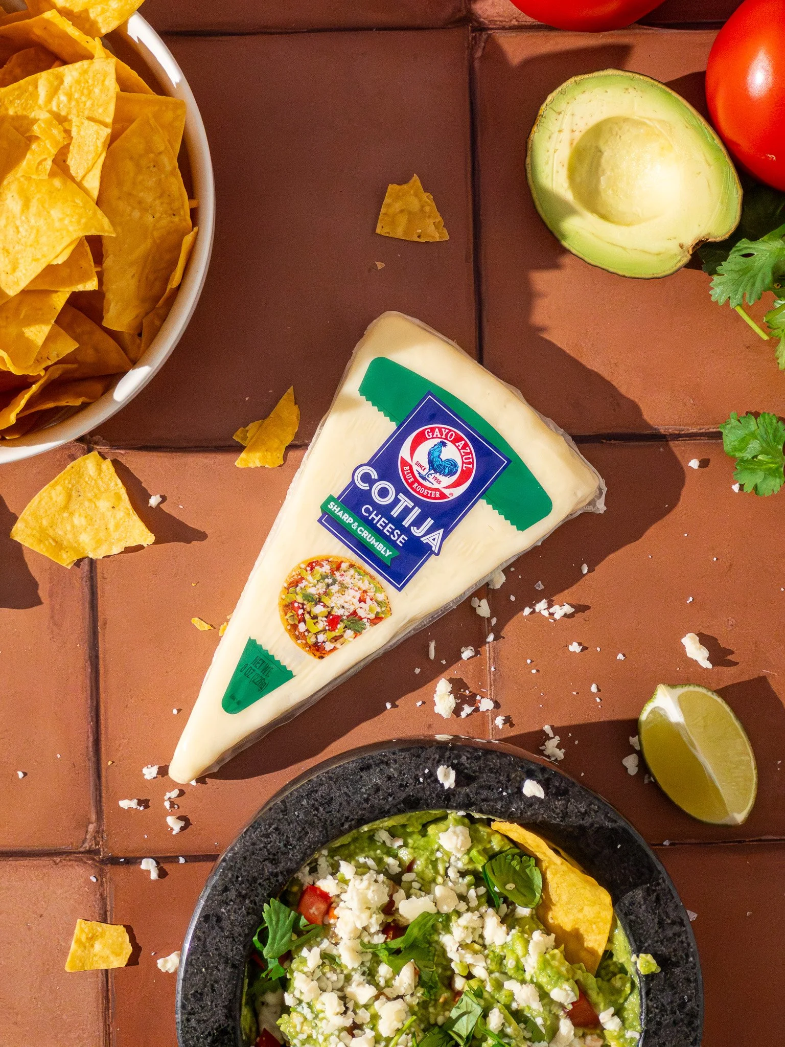 A top-down view of a food scene with fresh ingredients and dishes. There is a bowl of yellow tortilla chips, a wedge of cotija cheese, a half avocado, a lime wedge, tomatoes, cilantro, and a bowl of guacamole topped with crumbled cotija cheese.
