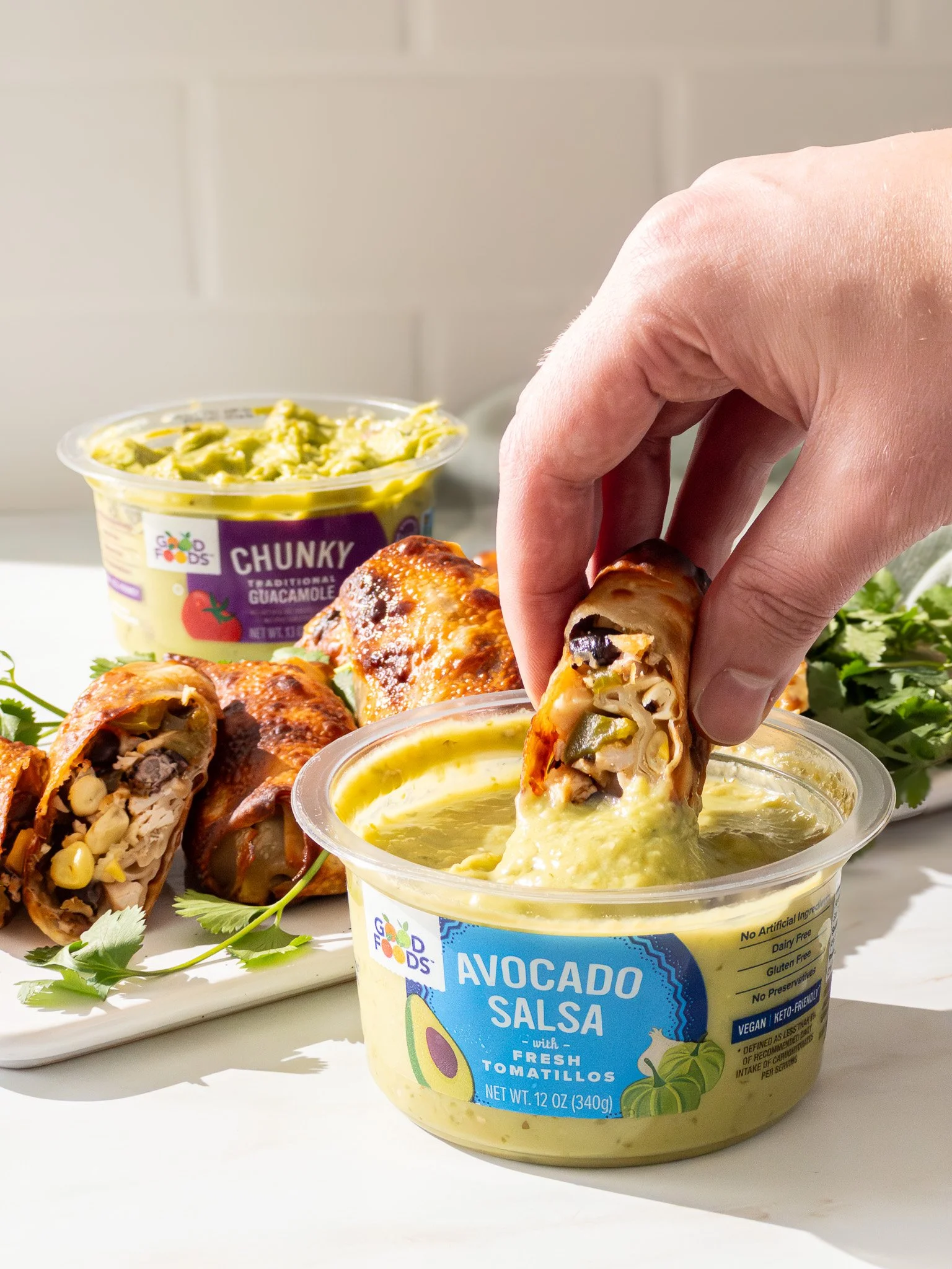A hand dipping a chicken and vegetable burrito into a cup of avocado salsa, with a plate of burritos and a container of guacamole in the background.