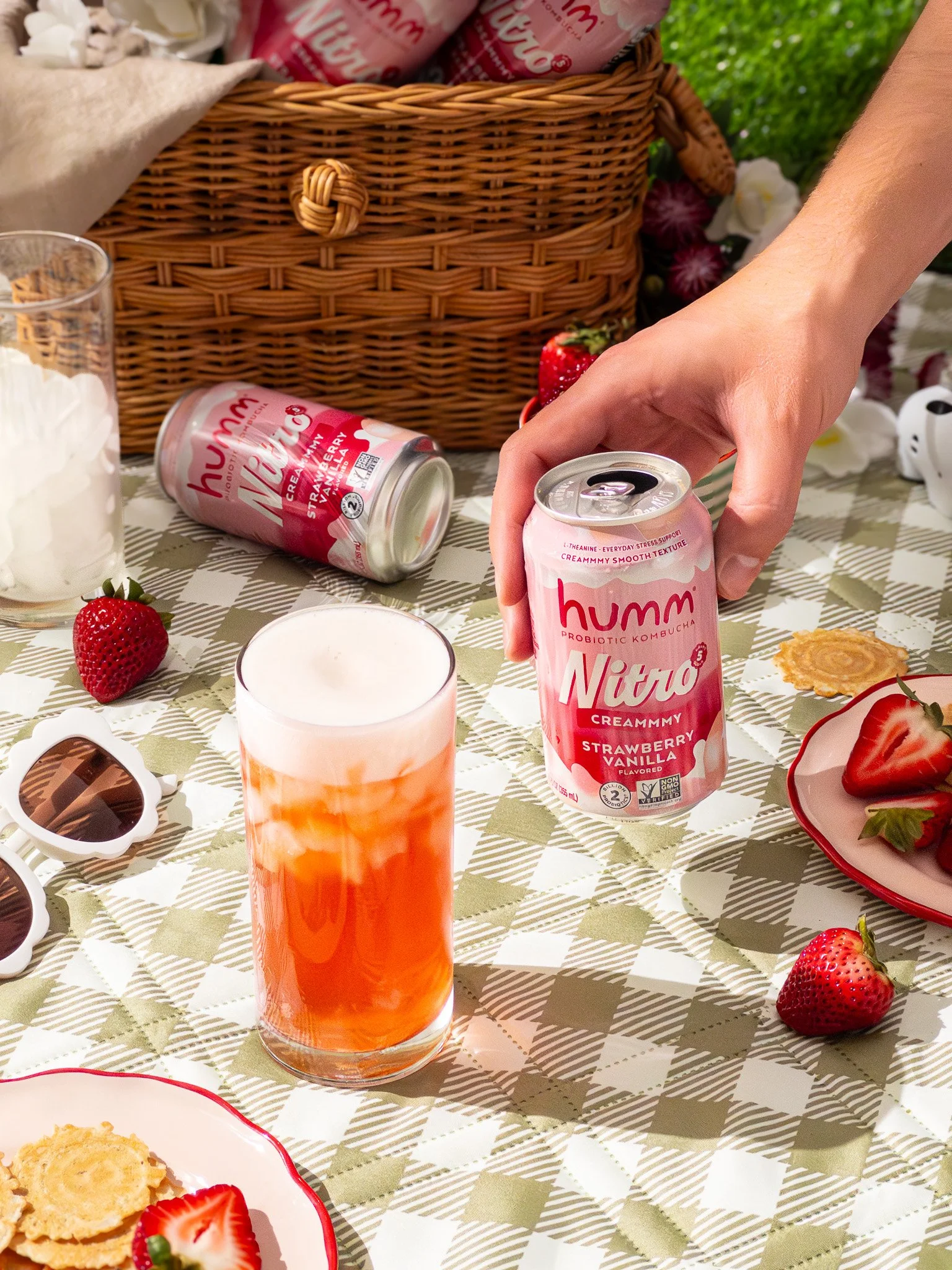A person holding a can of humm Nitro creammmy strawberry vanilla flavored probiotic kombucha on a table with strawberries, a glass of poured kombucha, cookies, and a wicker basket in the background.