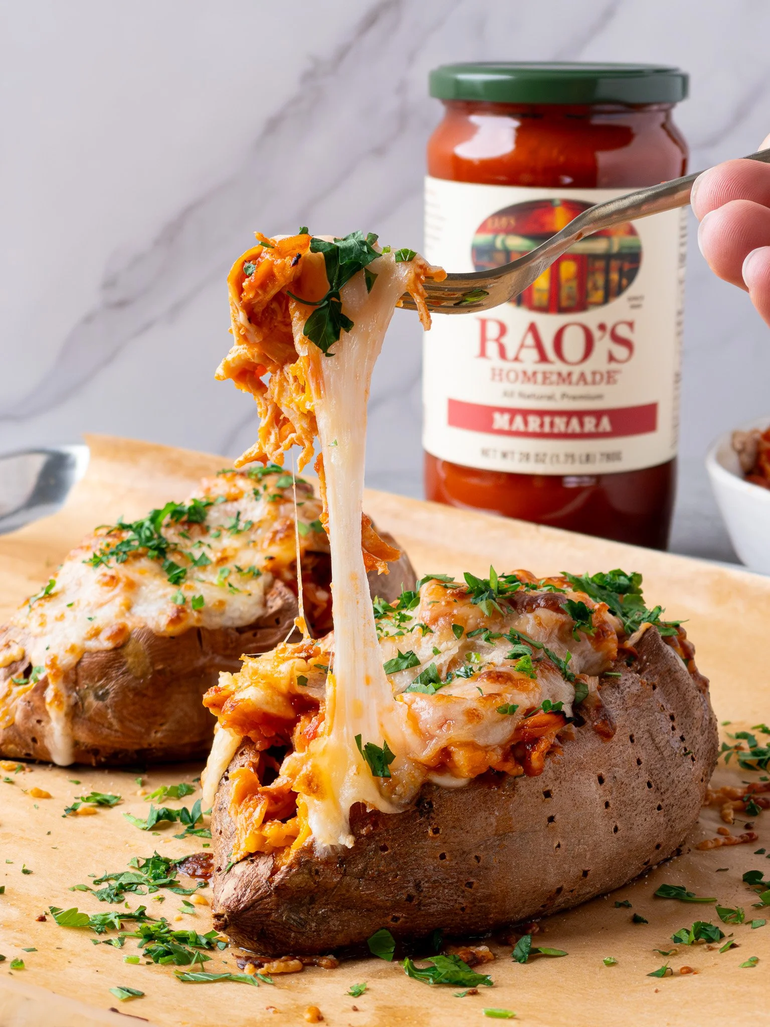 Baked sweet potato topped with cheesy chicken and marinara sauce, garnished with chopped parsley, with a jar of Rao's homemade marinara sauce in the background.
