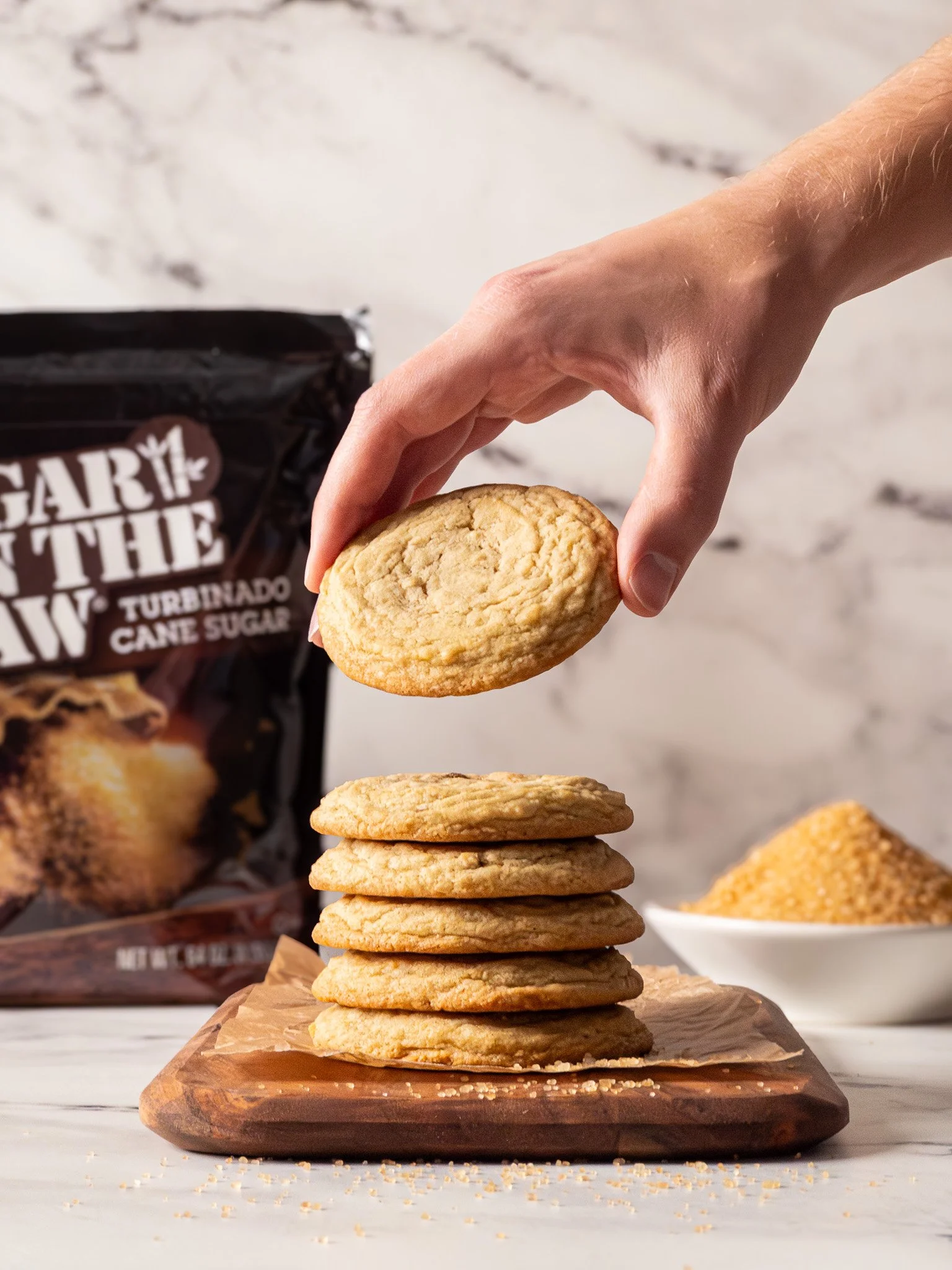 A hand holding a cookie above a stack of cookies on a wooden board, with a bag of caramel cane sugar and a bowl of yellow sugar in the background.