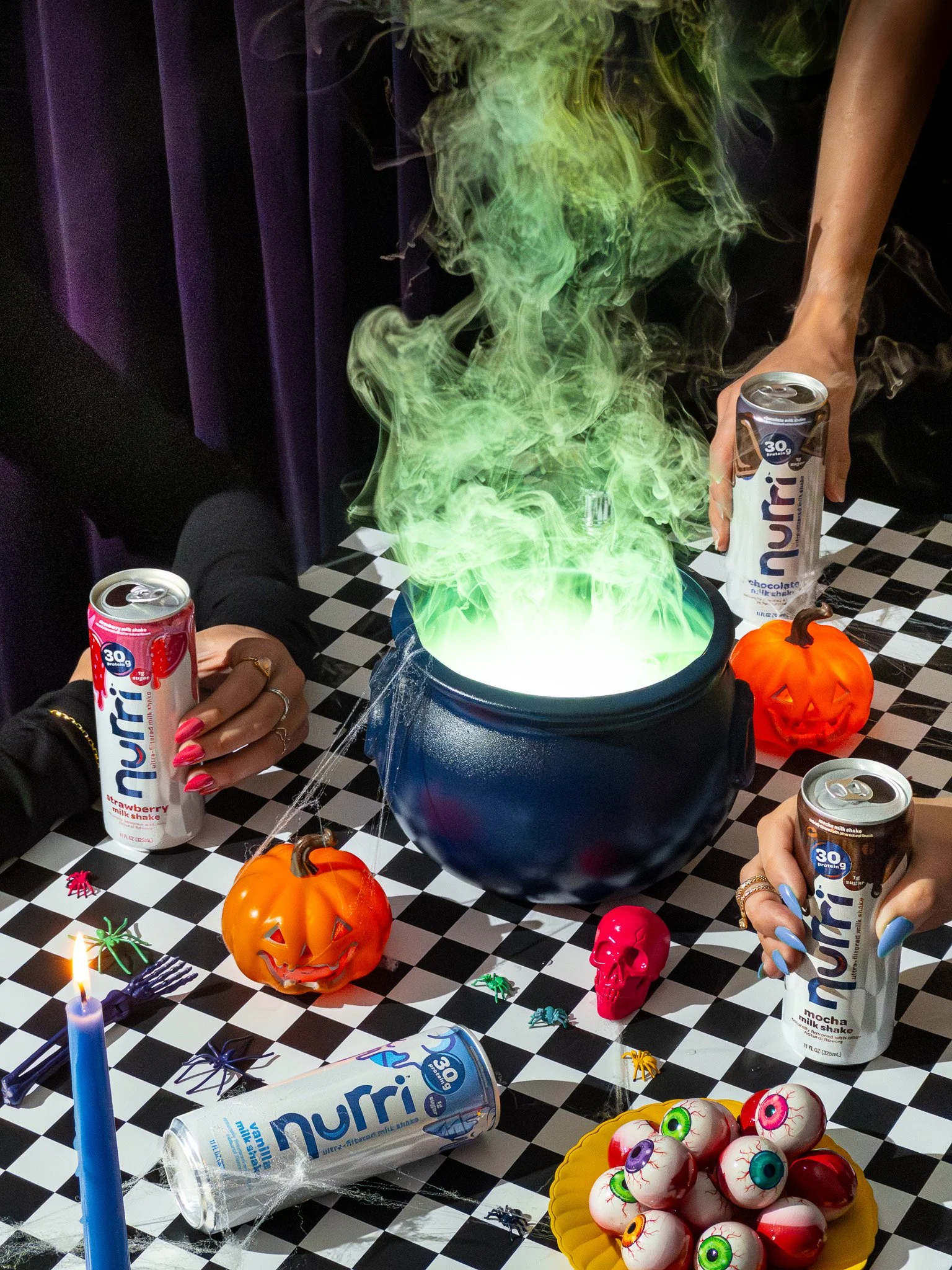 Halloween-themed party scene with a cauldron emitting green smoke, orange pumpkins, eyeball bowl, plastic spiders, skull, and people holding cans of NuRii drink.