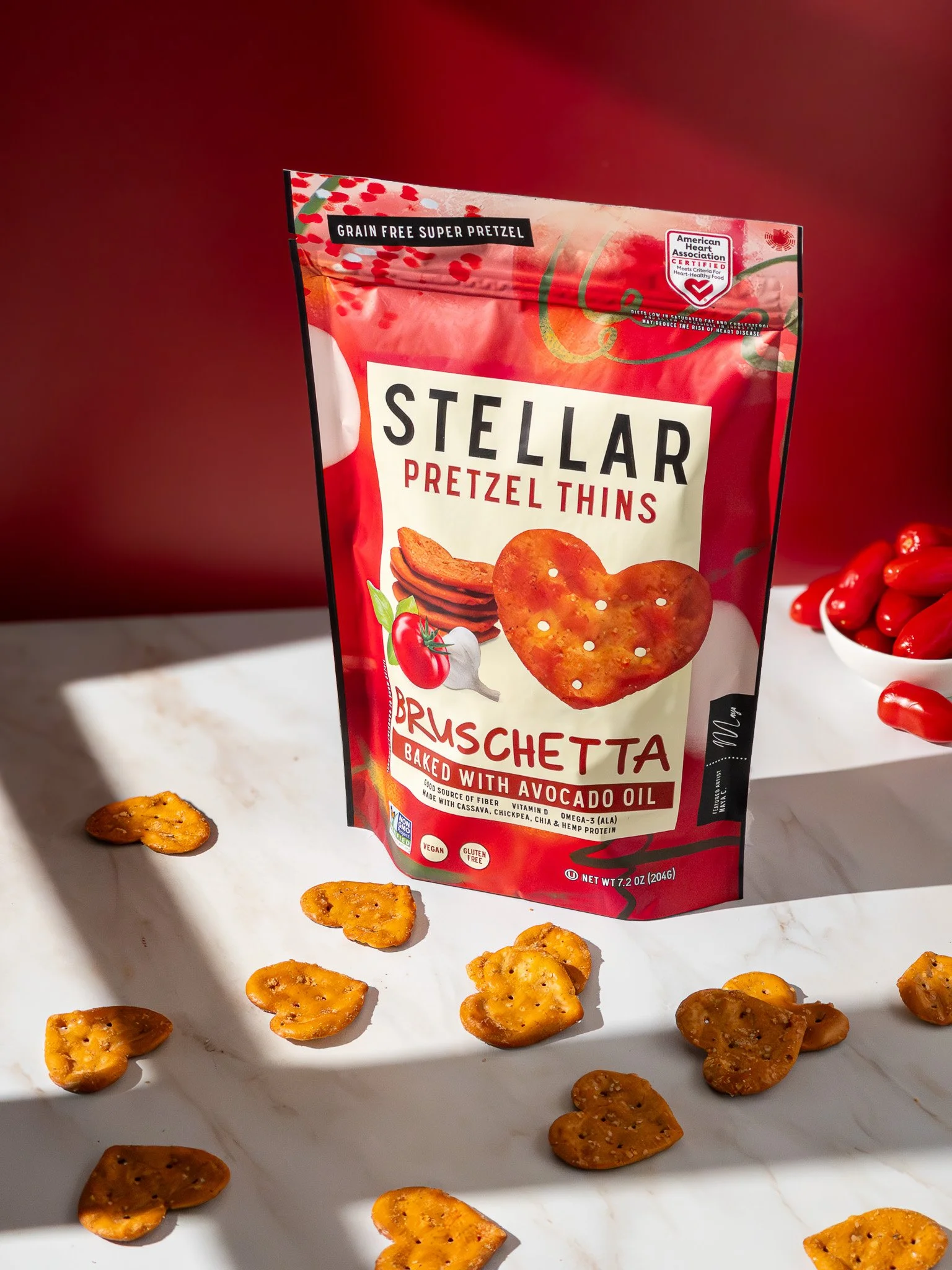 Package of Stellar Pretzel Thins Bruschetta baked with avocado oil on a white surface with scattered heart-shaped pretzel snacks and a bowl of cherry tomatoes in the background.