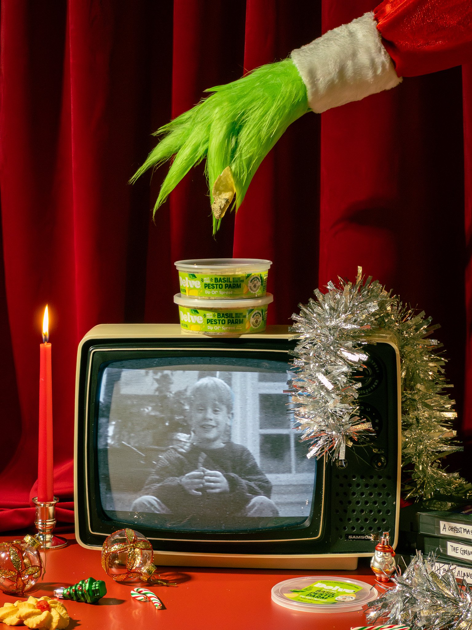 Retro TV displaying a black-and-white photo of a young boy giving a thumbs-up, surrounded by Christmas decorations including ornaments, tinsel, a candle, and snack plates. A green Dr. Oetker pesto container is on top of the TV, and a large green furr
