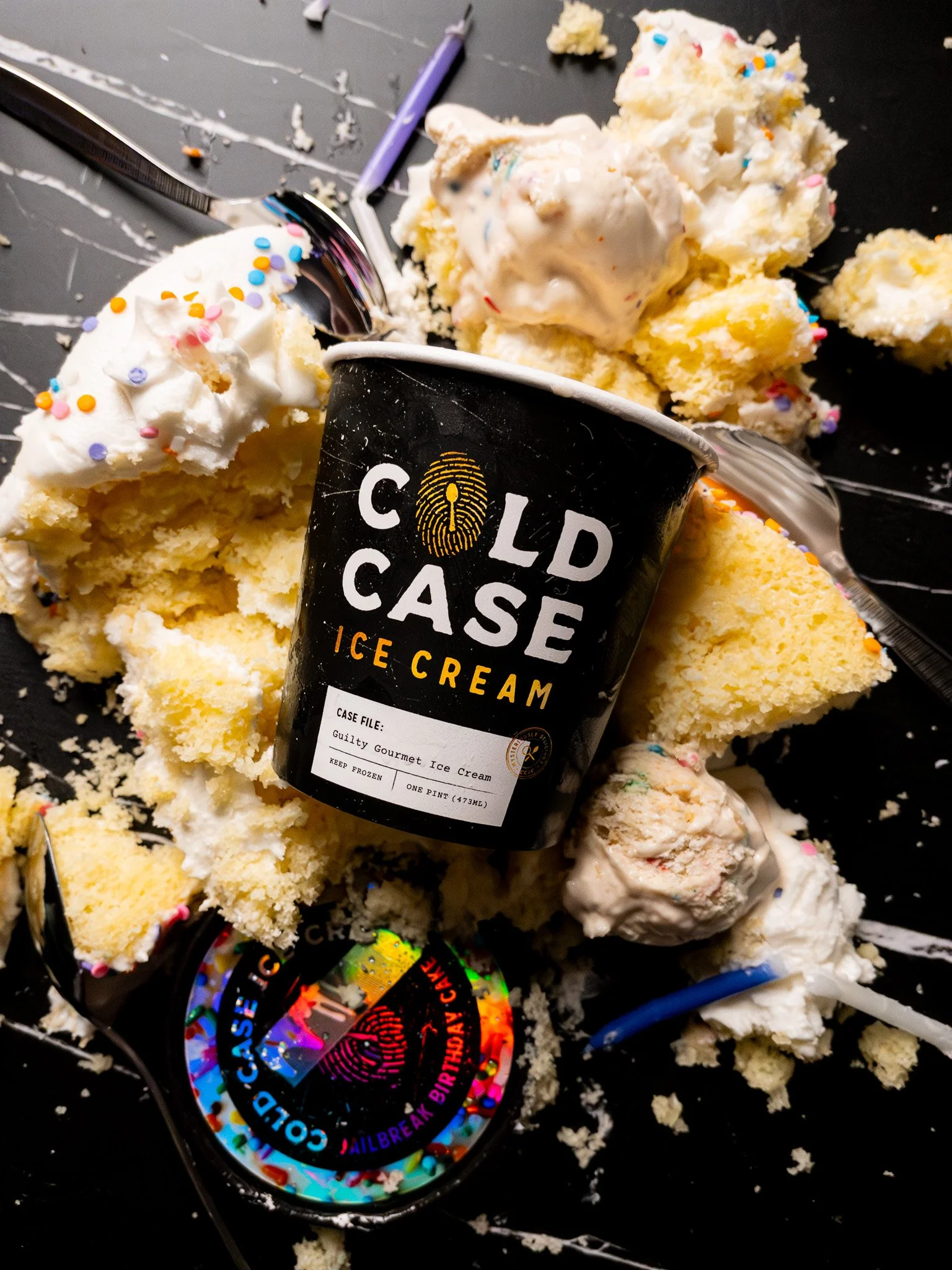 Scoops of vanilla ice cream with colorful sprinkles and ice cream crumbs scattered on a black surface, with a Cold Case Ice Cream pint container and a holographic sticker celebrating a birthday.
