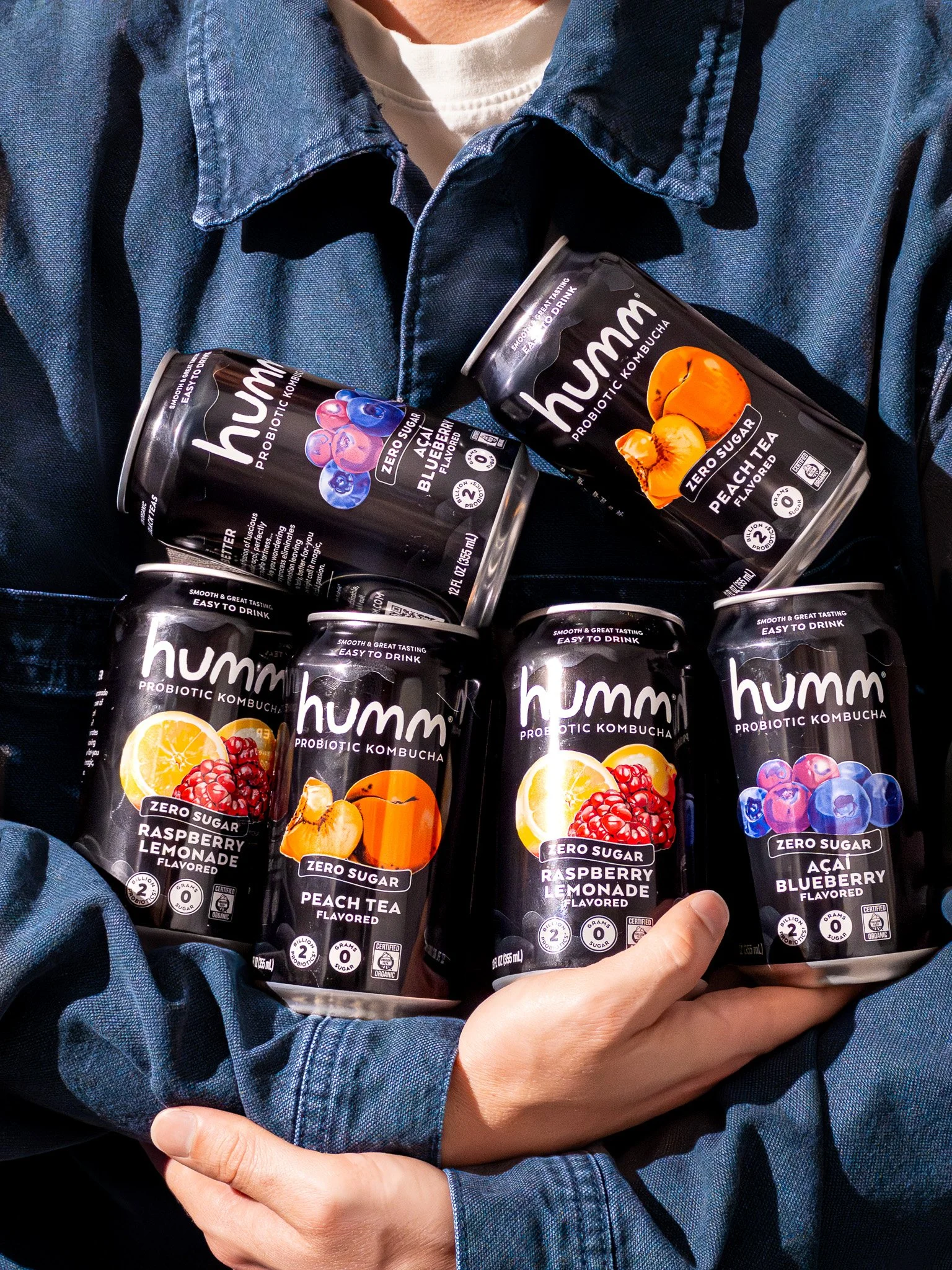 A person in a denim jacket holding six cans of Humm probiotic drinks, with flavors including raspberry lemonade, peach tea, and acai blueberry.