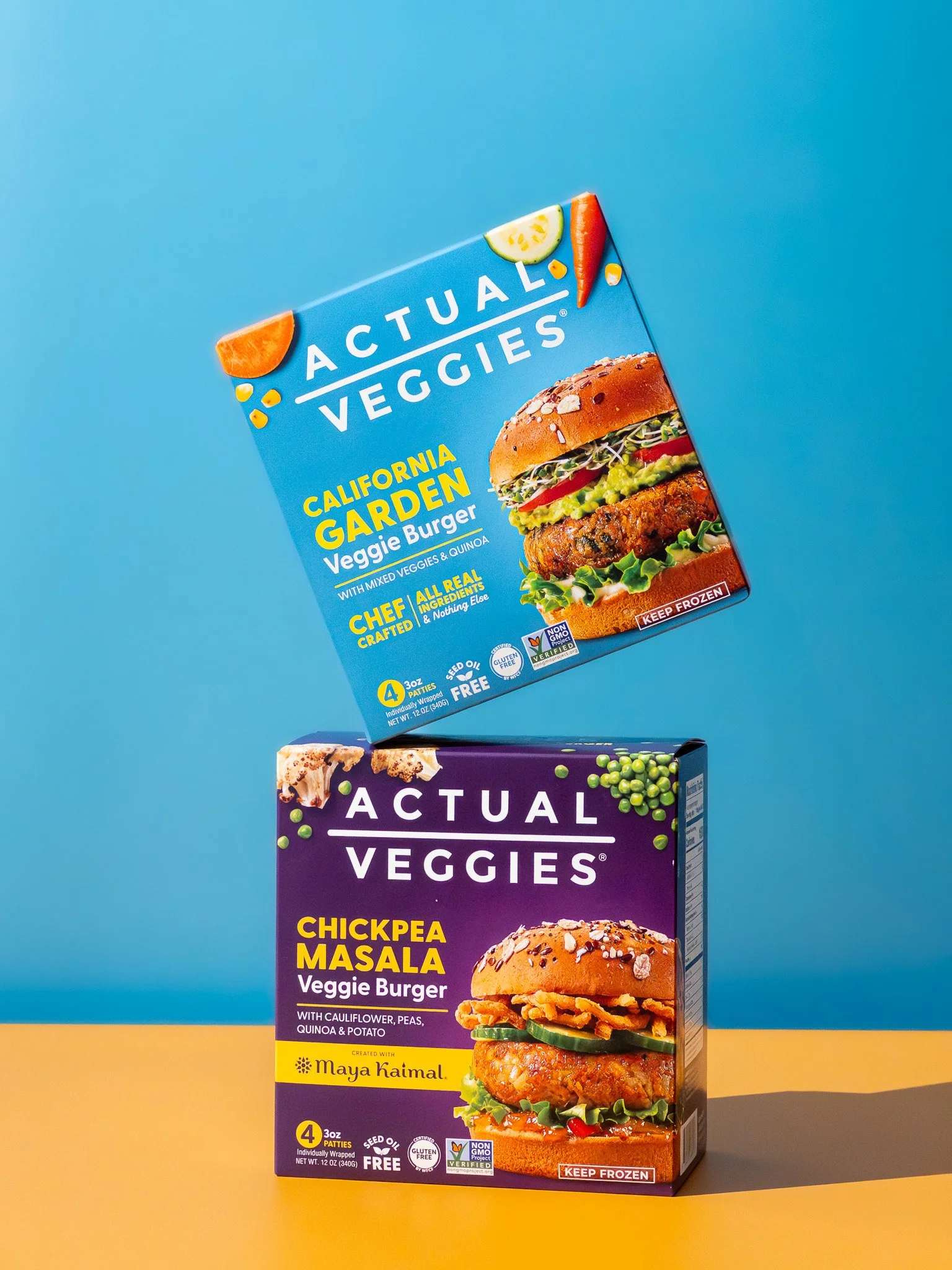 Two boxes of Actual Veggies veggie burger patties, one blue box labeled California Garden Veggie Burger with assorted vegetables and quinoa, and one purple box labeled Chickpea Masala Veggie Burger with cauliflower, peas, quinoa, and potato, against 