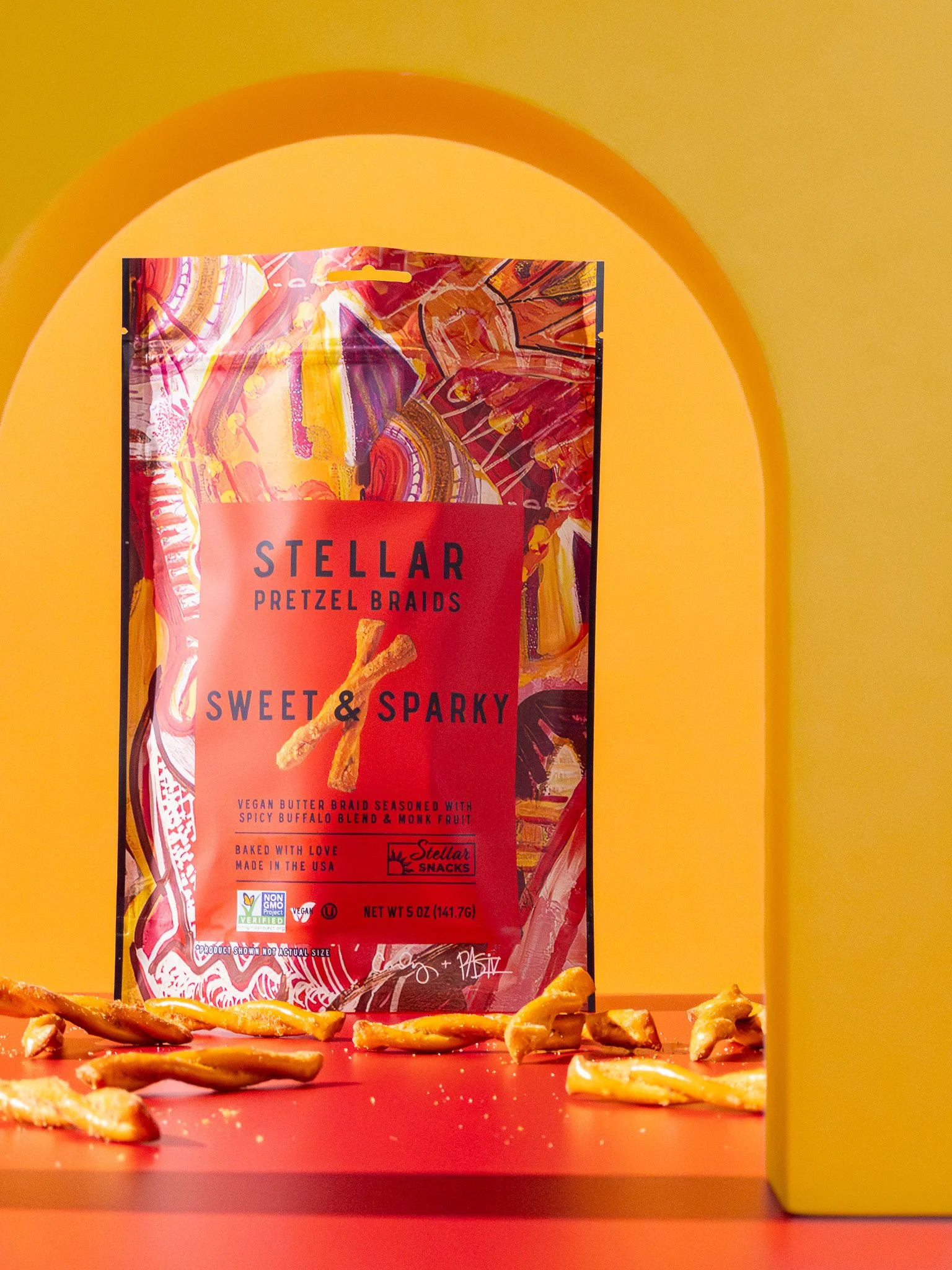 A bag of Stellar Pretzel Braids with the label reading 'Sweet & Sparky' placed on a red surface with some pretzels scattered around, framed by a yellow arch-shaped structure against a yellow background.