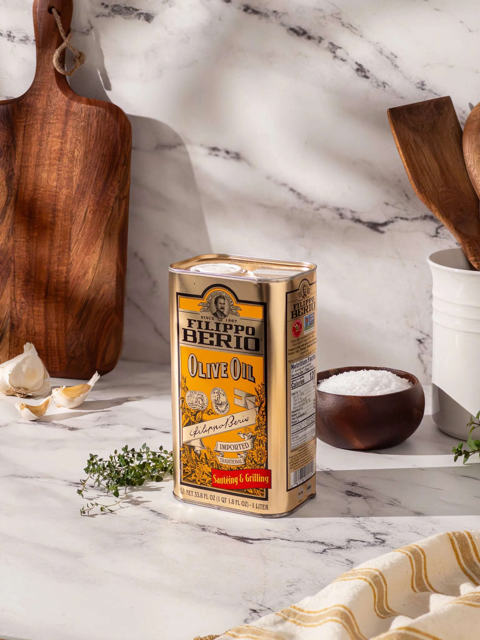 A can of Filippo Berio Olive Oil on a marble countertop with wooden cutting boards, a small white bowl of salt, and garlic cloves.