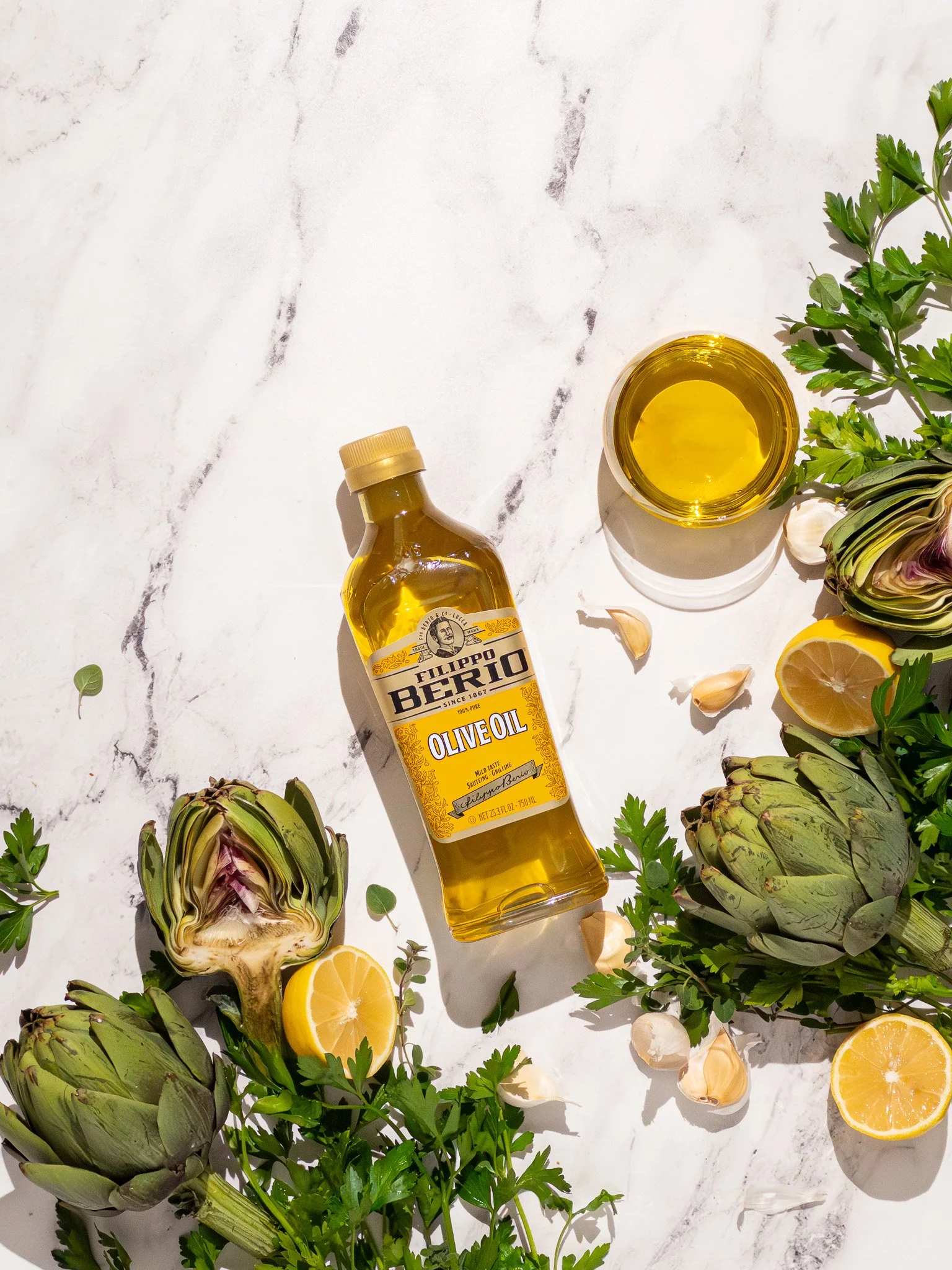 Bottle of Filippo Berio olive oil, halved artichokes, lemon halves, garlic cloves, and fresh parsley on a white marble surface.