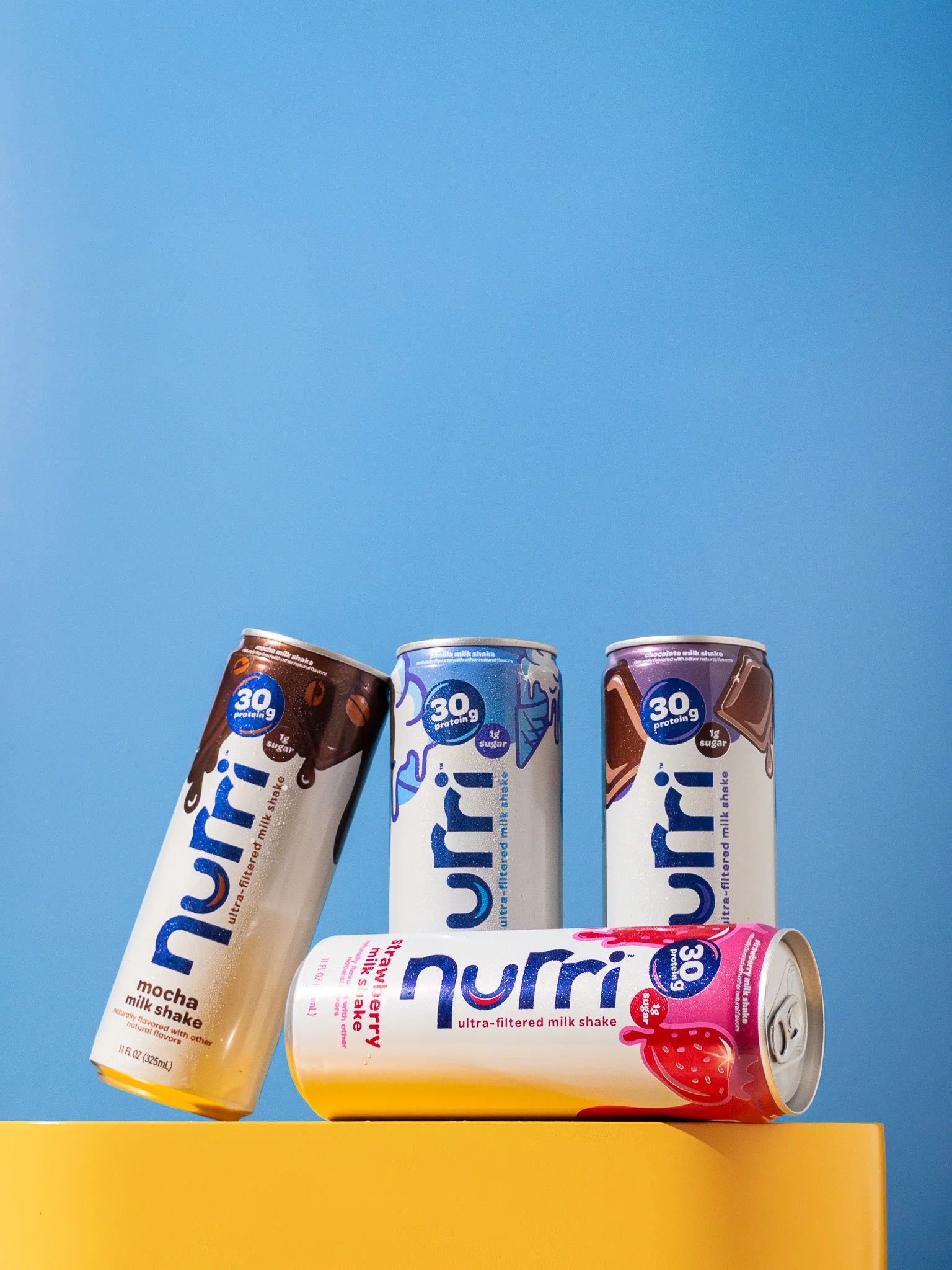 Four Nu Rri milkshake cans in various flavors, including mocha, vanilla, strawberry, and chocolate, are displayed on a yellow surface against a light blue background.