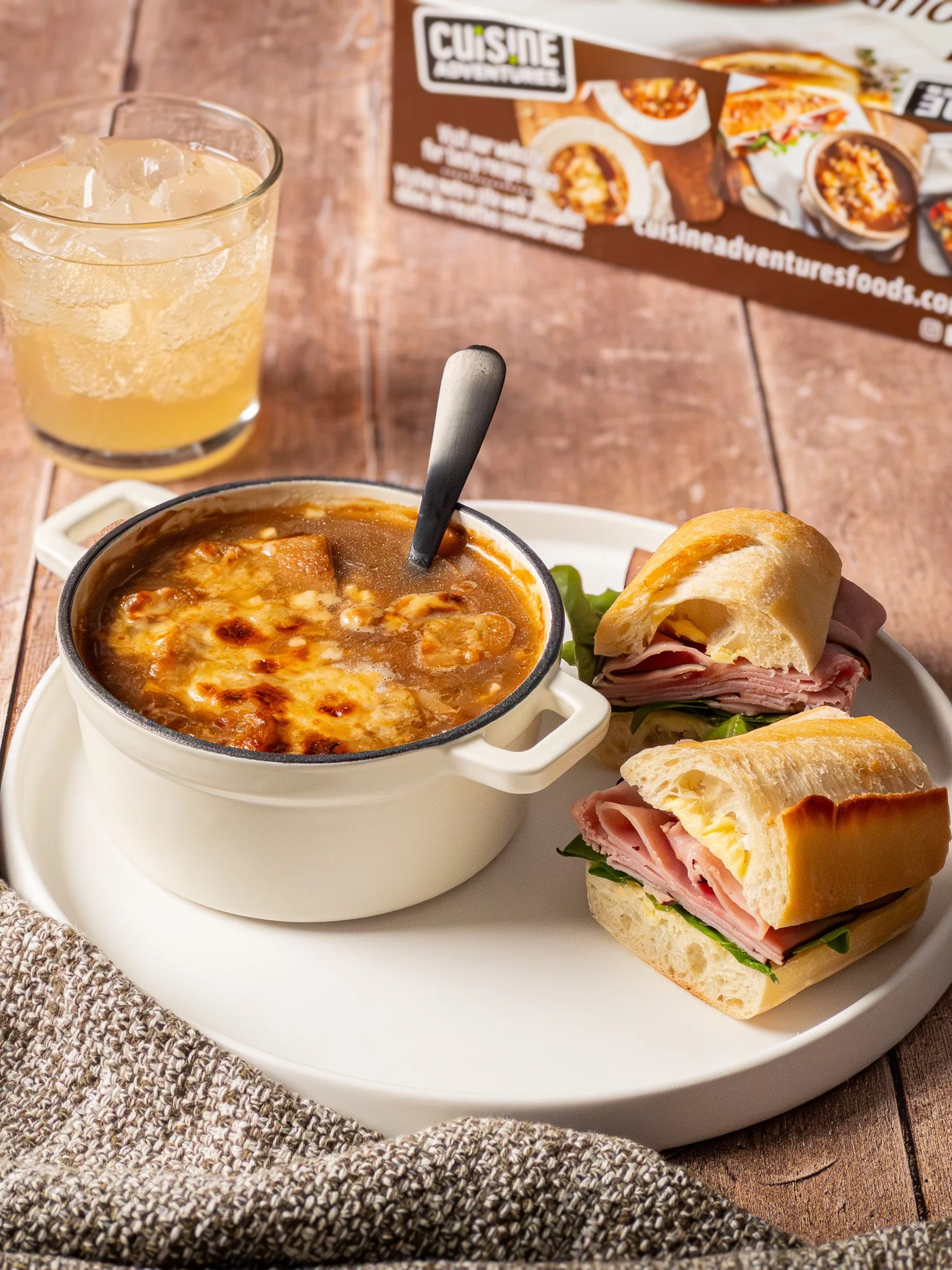 A wooden table with a white plate holding two sandwich wraps, a bowl of French onion soup with a spoon, and a glass of iced apple juice. A menu is in the background.