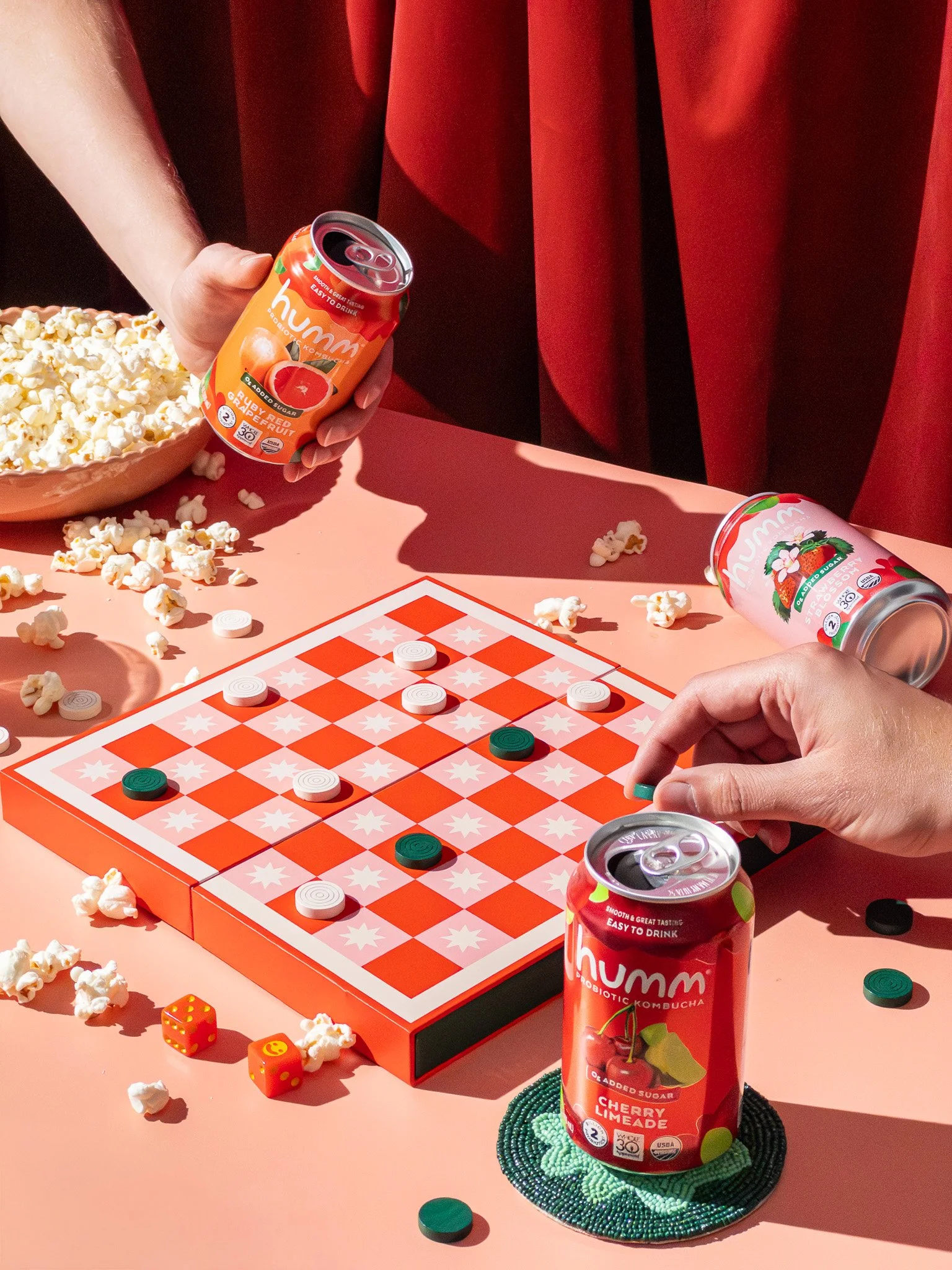 Two people playing checkers on a red and white checkered board, with popcorn, two cans of cherry limeade, and two small dice on a pink table, with a red curtain in the background.