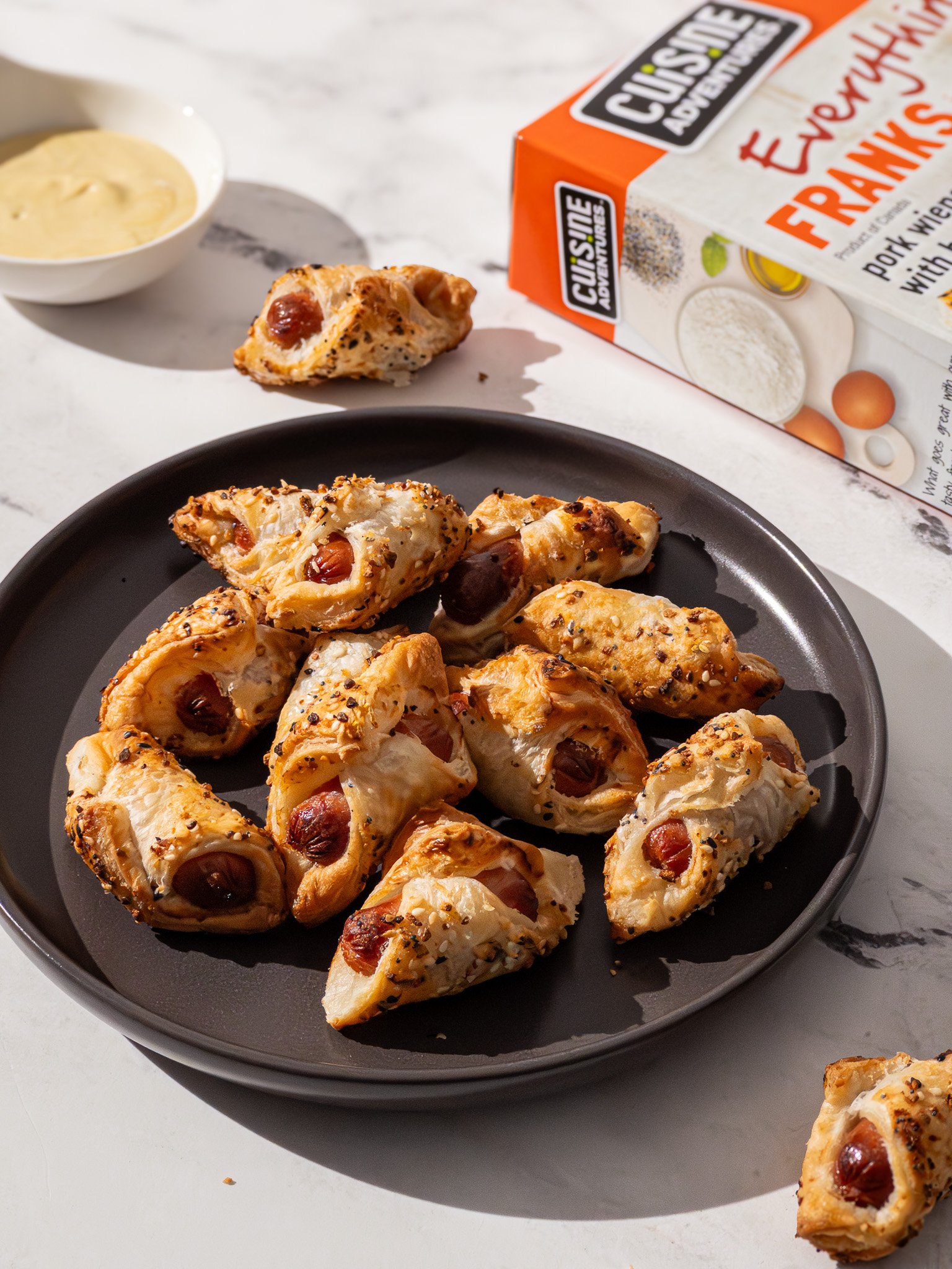 Plate of stuffed pastry bites with sausage and cheese, served on a black plate with a dipping sauce, with a box of Crackin' Adventure Everything Breakfast mix in the background.