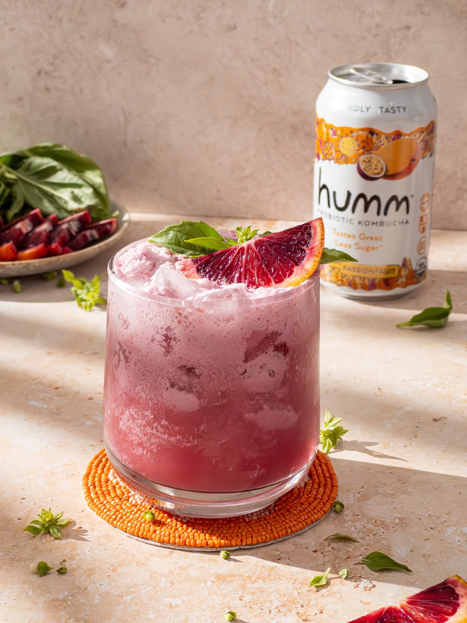 A pink beverage with ice, garnished with a slice of blood orange and fresh basil, placed on an orange beaded coaster. A can of Humm probiotic kombucha is in the background, along with a plate of sliced blood orange and greens.