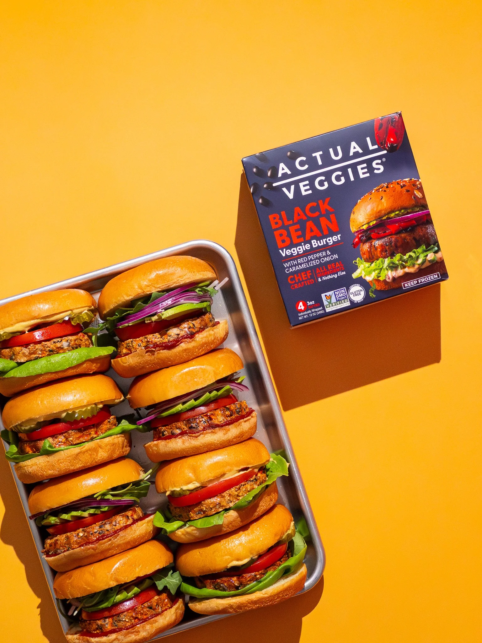 Tray of multiple veggie burgers with lettuce, tomato, onion, and a patty, next to a box of Aactua veggie's Black Bean veggie burger on a yellow background.