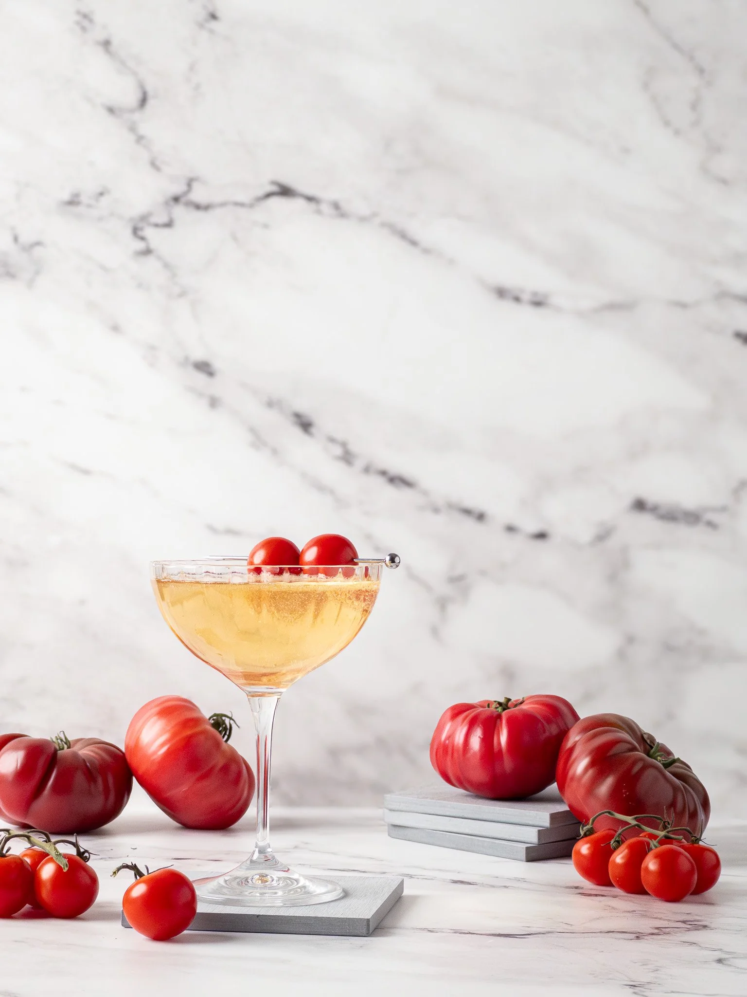 A cocktail glass with yellowish liquid and tomatoes on top, surrounded by various ripe tomatoes on a white surface with a marble background.