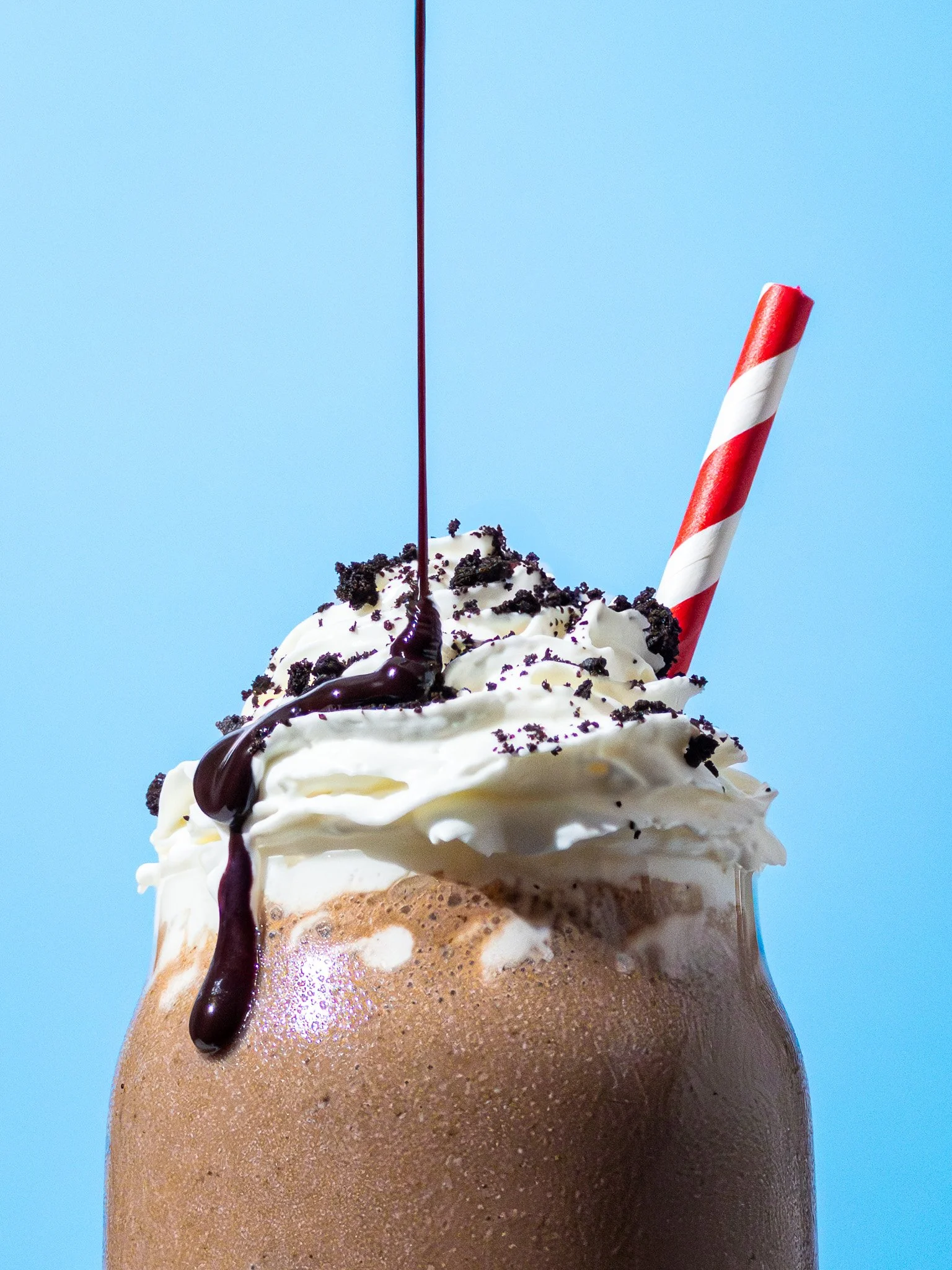 Chocolate milkshake topped with whipped cream, chocolate syrup, cookie crumbles, and a red-and-white striped straw on a light blue background.