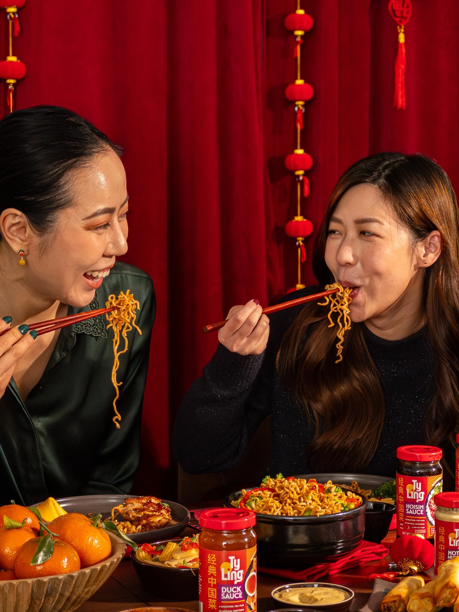 Two women sharing a meal of noodles, laughing and enjoying each other's company at a table decorated for a Chinese celebration, with red lanterns and jars of Asian sauce.