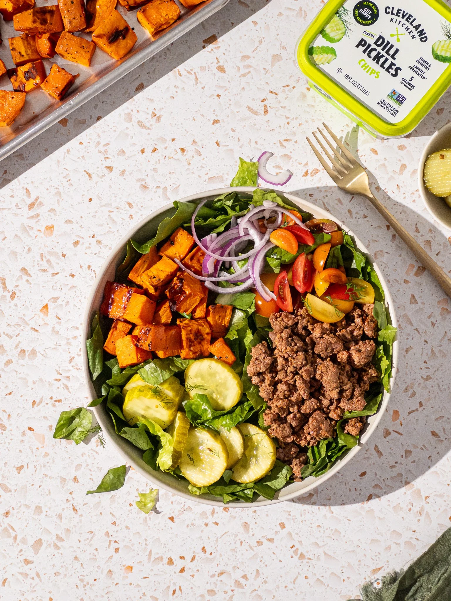 A bowl of salad containing grilled sweet potatoes, ground beef, sliced cucumbers, cherry tomatoes, and sliced red onions, on a speckled white surface.