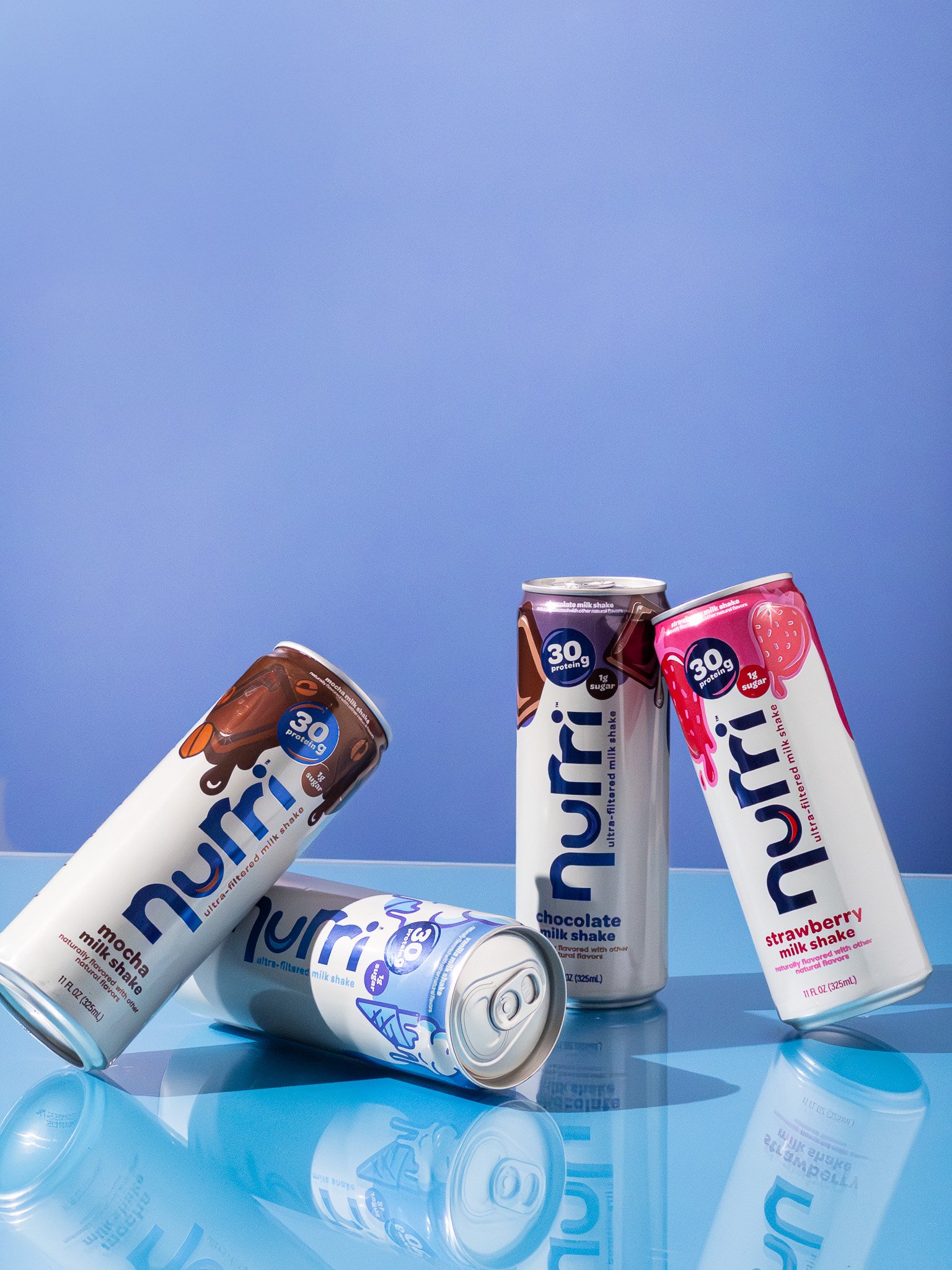 Assorted cans of Nirí milkshakes in various flavors including mocha, chocolate, and strawberry, placed on a reflective surface against a blue background.