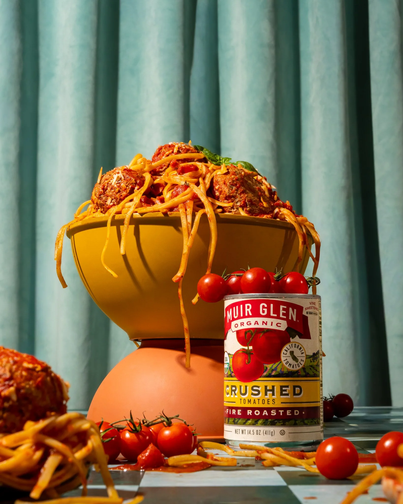 A bowl of spaghetti with meatballs, cherry tomatoes, and basil on a pedestal, with a can of crushed tomatoes and scattered tomatoes and pasta around.
