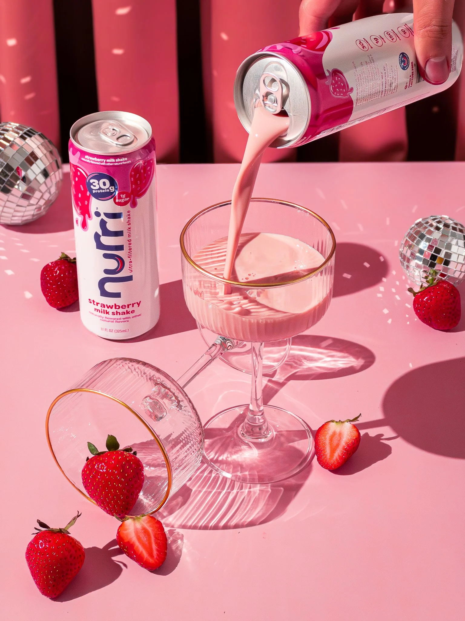 A can of strawberry milk shake pouring into a glass with strawberries scattered on a pink surface. Two disco balls are also visible on the table.