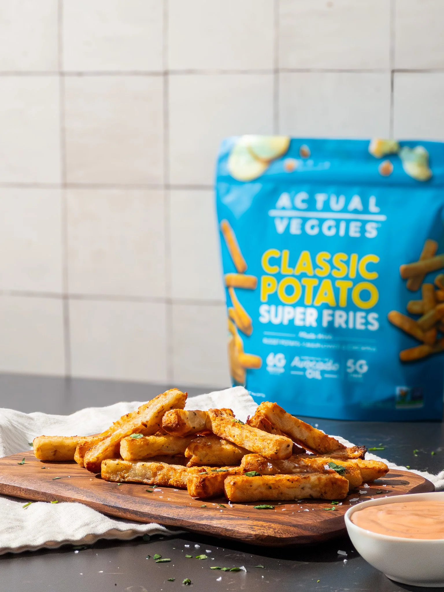 A wooden cutting board with seasoned French fries on it, served with a small bowl of dipping sauce, in front of a blue bag of actual Veggies classic potato super fries, on a black countertop against a tiled wall background.