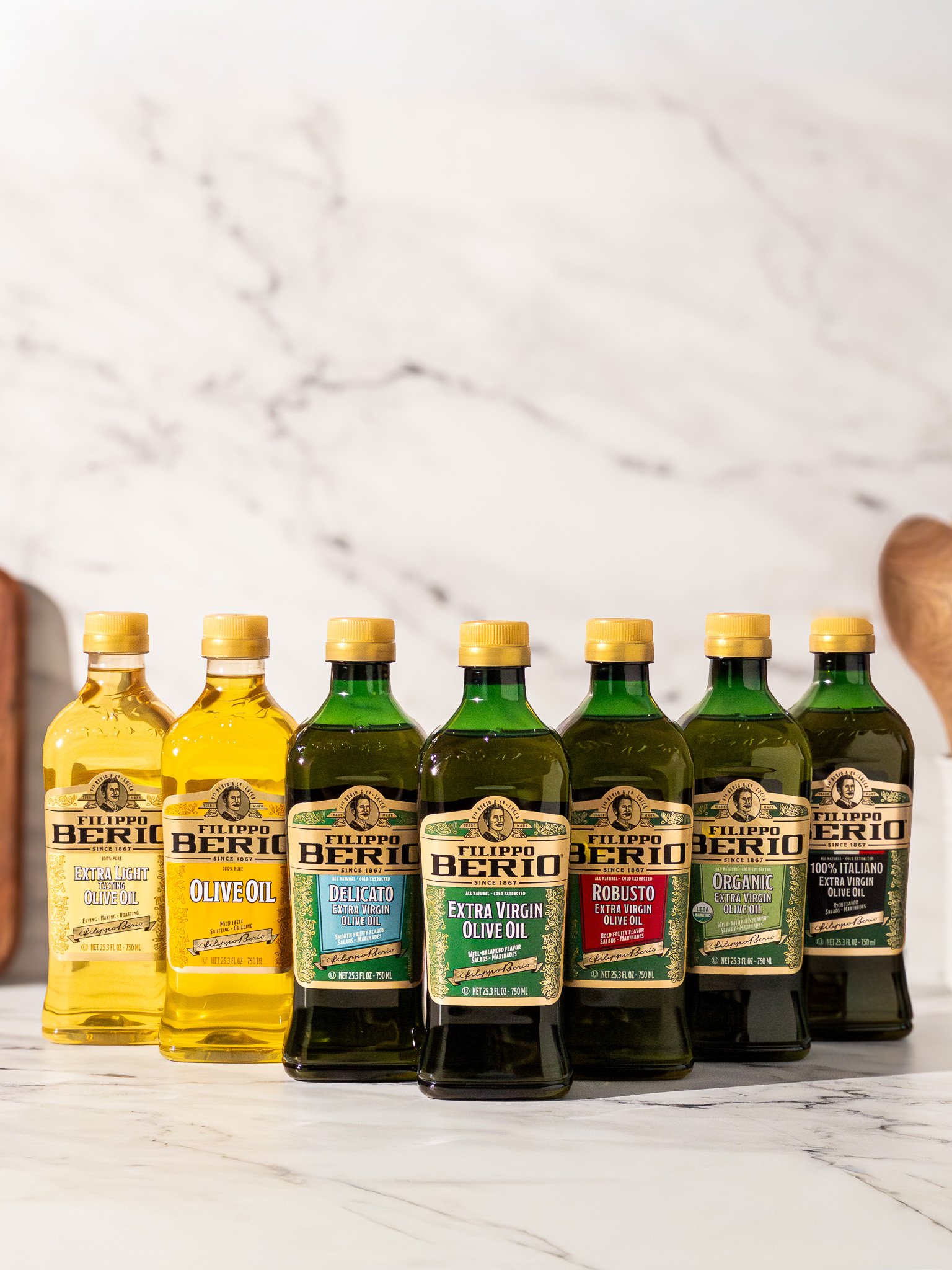 Six bottles of Filippo Berio olive oils on a marble surface, with various types including extra virgin, organic, and extra light.