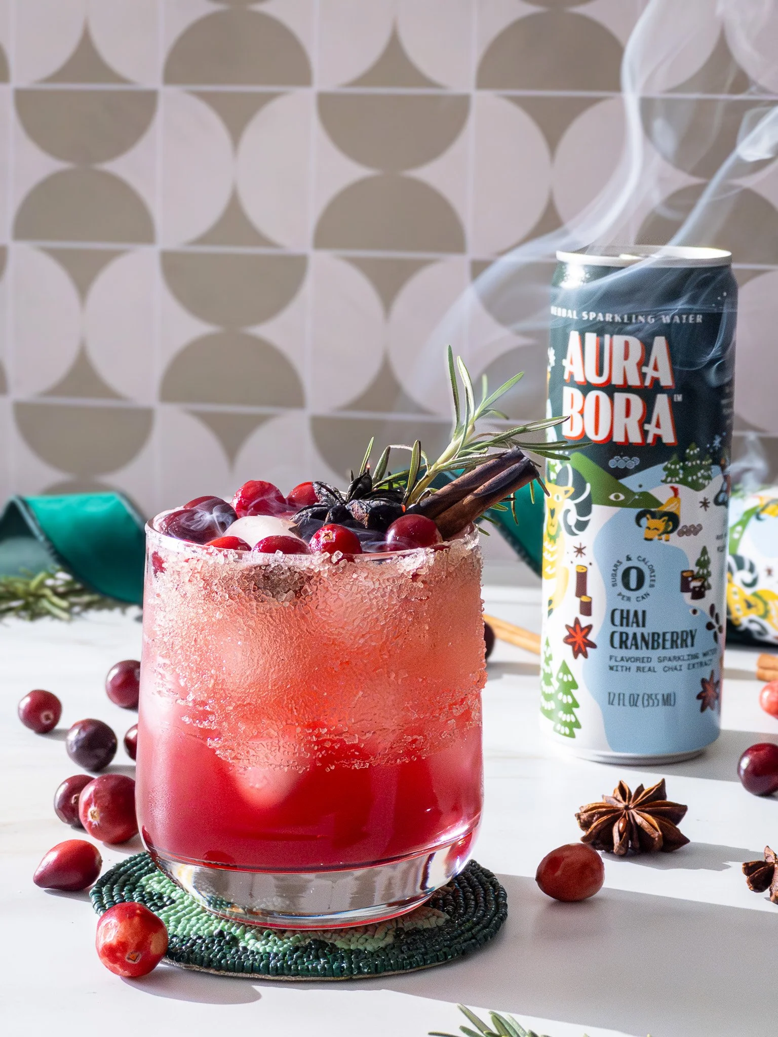 A glass with a pink-red beverage, ice, cranberries, star anise, cinnamon stick, and rosemary, placed on a woven coaster. Behind it is a can of Aura Bora Chai Cranberry sparkling water, surrounded by scattered cranberries, star anise, and rosemary, on
