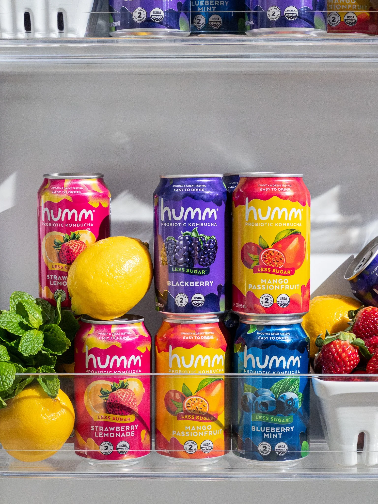 A display of canned probiotic kombucha drinks in various flavors, including strawberry lemonade, blackberry, mango passion fruit, and blueberry mint, with lemons and strawberries as decoration.