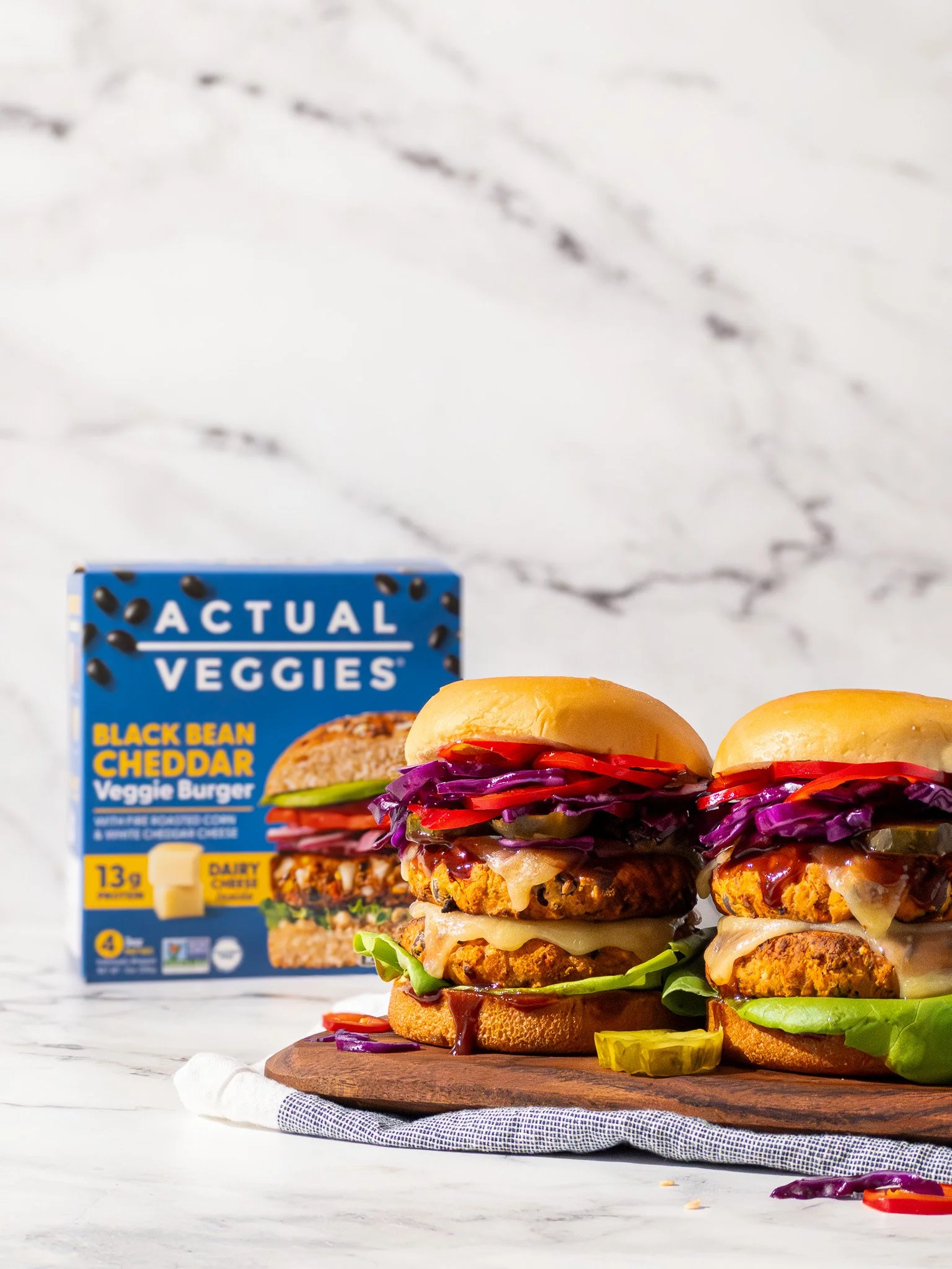 Two veggie burgers with lettuce, tomato, pickles, purple cabbage, and cheese on a wooden board, with a box of vegan burger patties in the background.