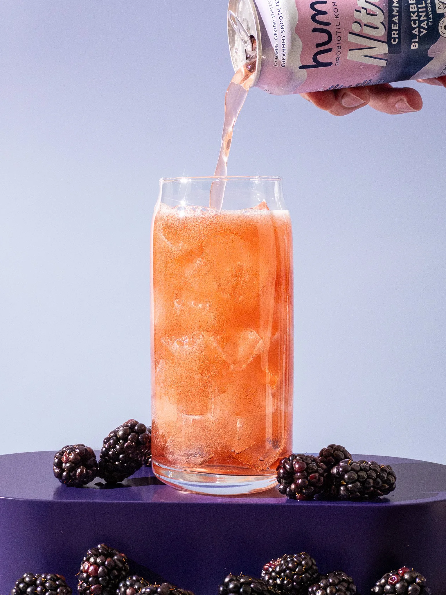 A hand pouring pink blackberry probiotic drink into a glass with ice, blackberries on purple surface, and a light background.