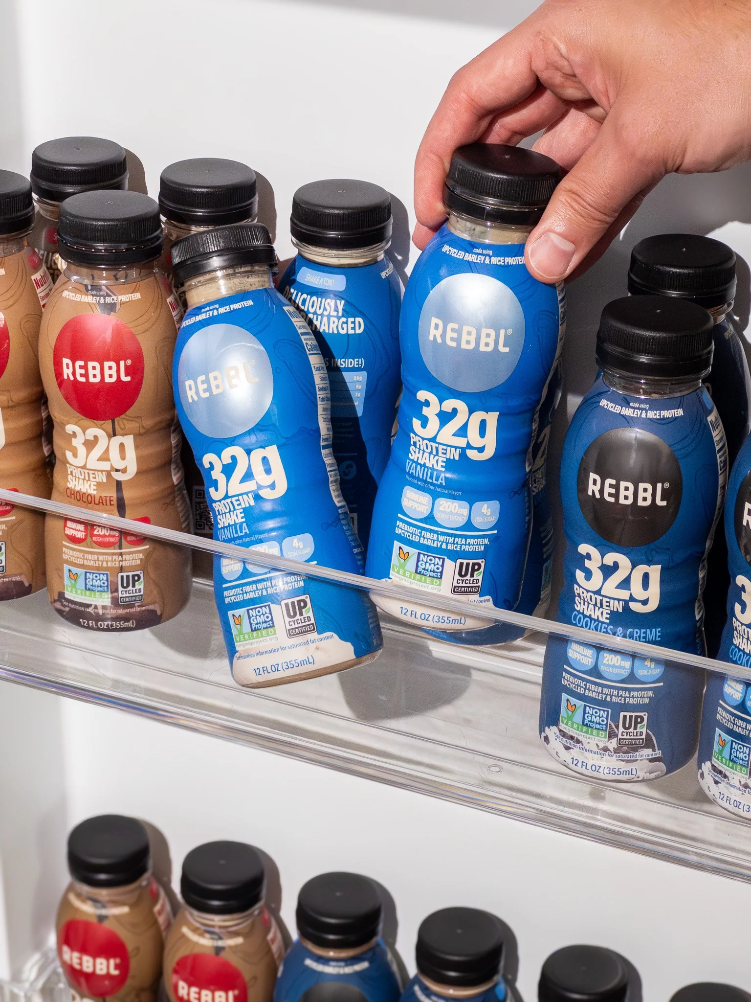 A person's hand reaching for a blue bottle of Rebbl vanilla protein shake on a store shelf, with other Rebbl protein shakes around it.